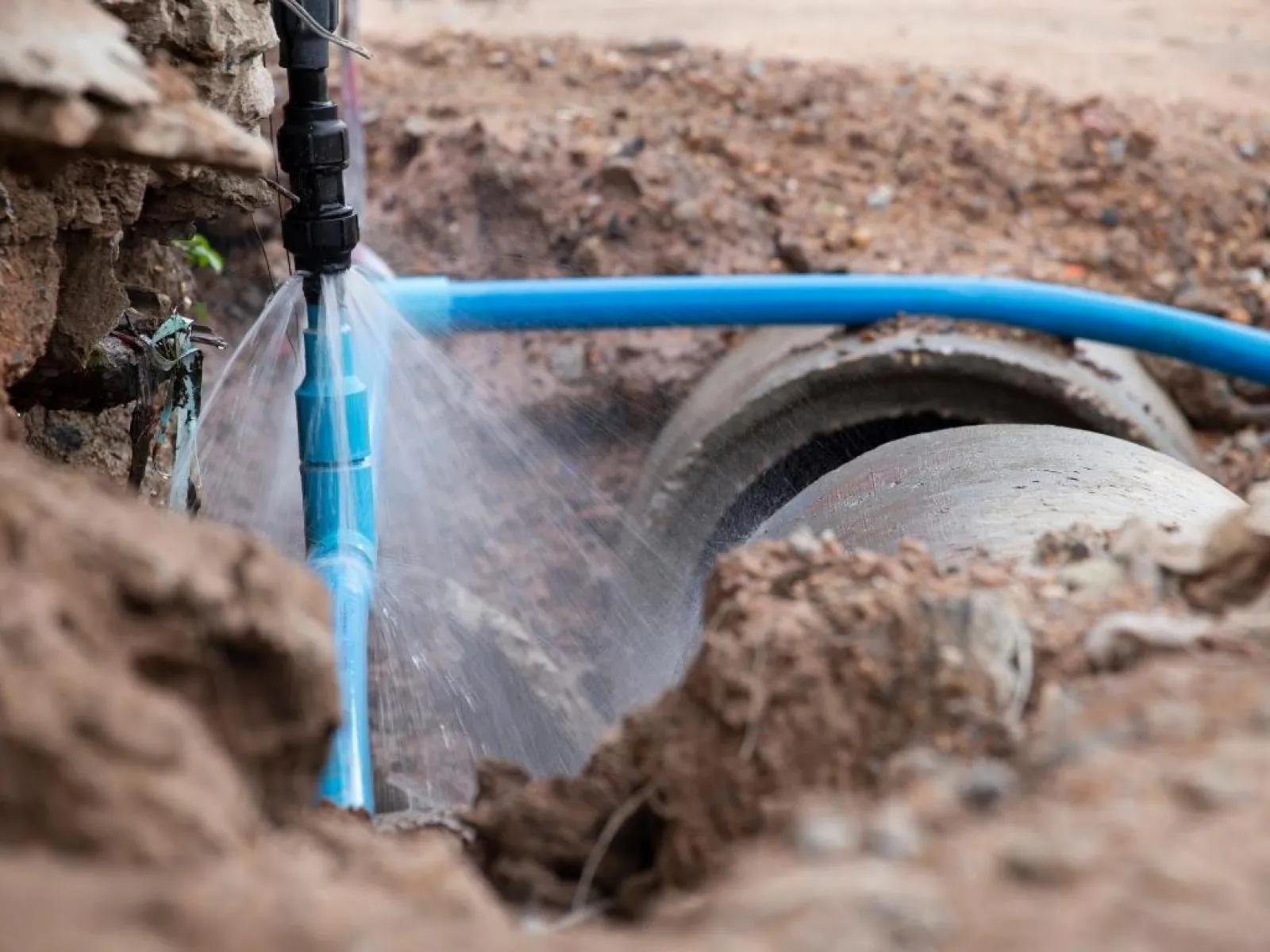 Leak in blue underground water pipe spraying water in a muddy excavation site during repairs.
