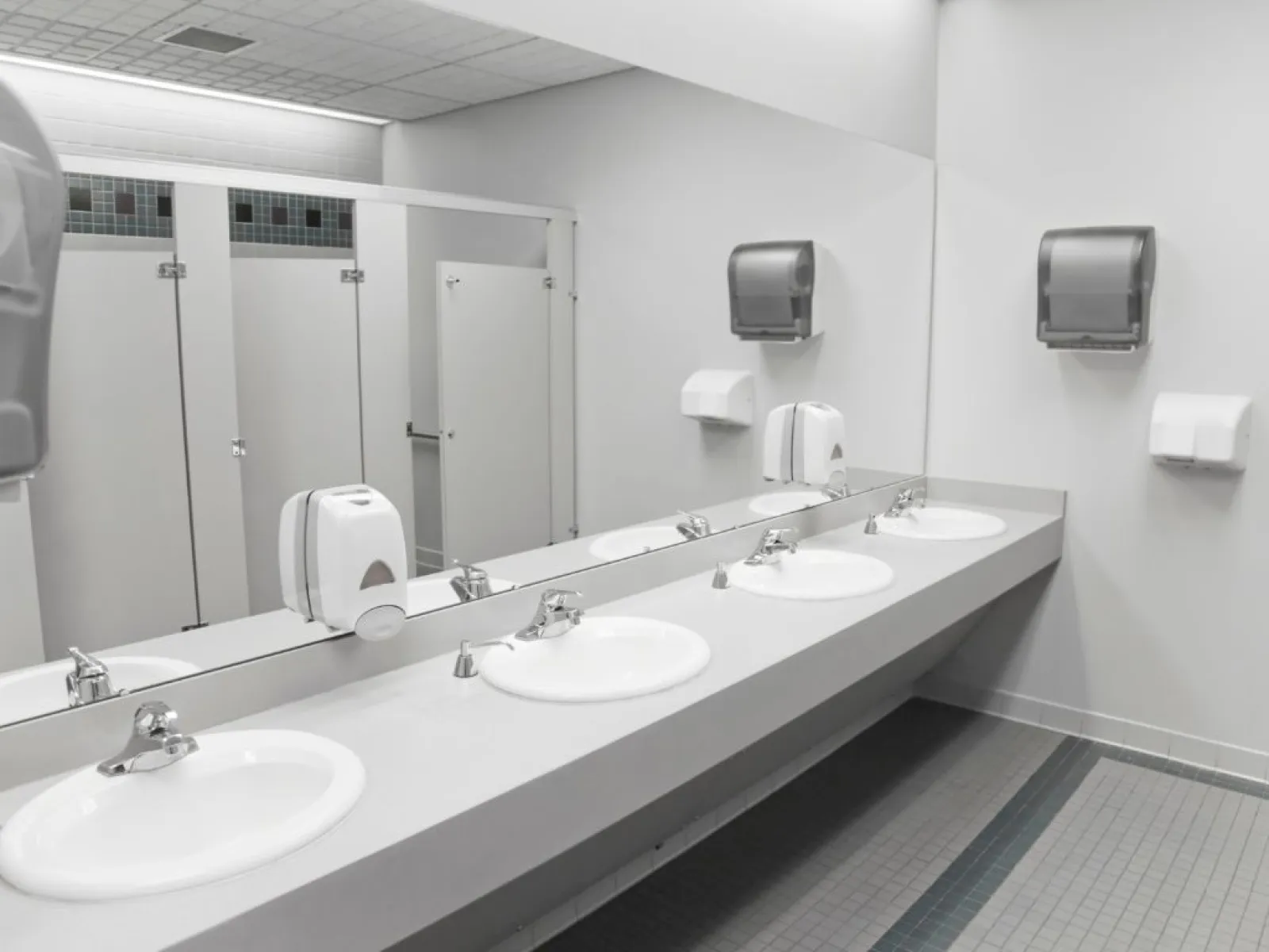 Clean public restroom with white sinks, large mirror, hand soap dispensers, paper towel holders, and toilet stalls.