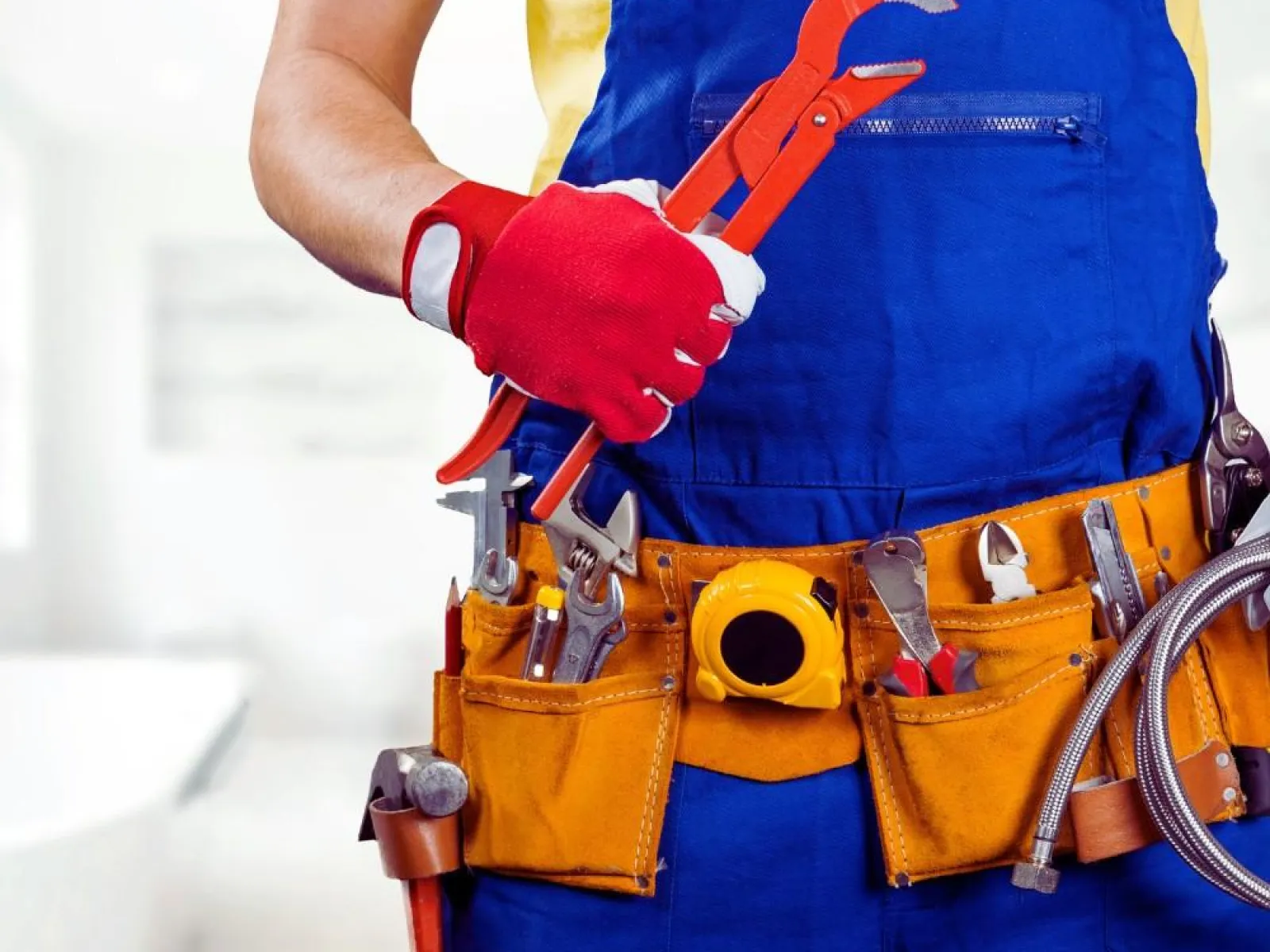 Plumber wearing blue overalls and red gloves holding a pipe wrench with a tool belt full of plumbing tools.