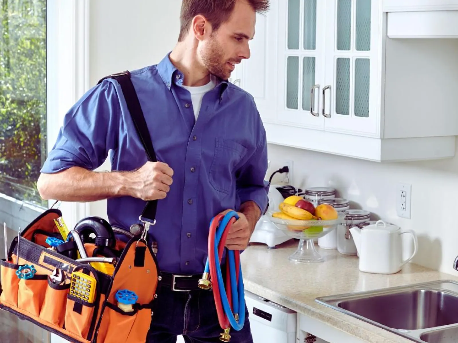 Plumber with tool bag and hoses inspecting kitchen sink area in a bright modern kitchen.