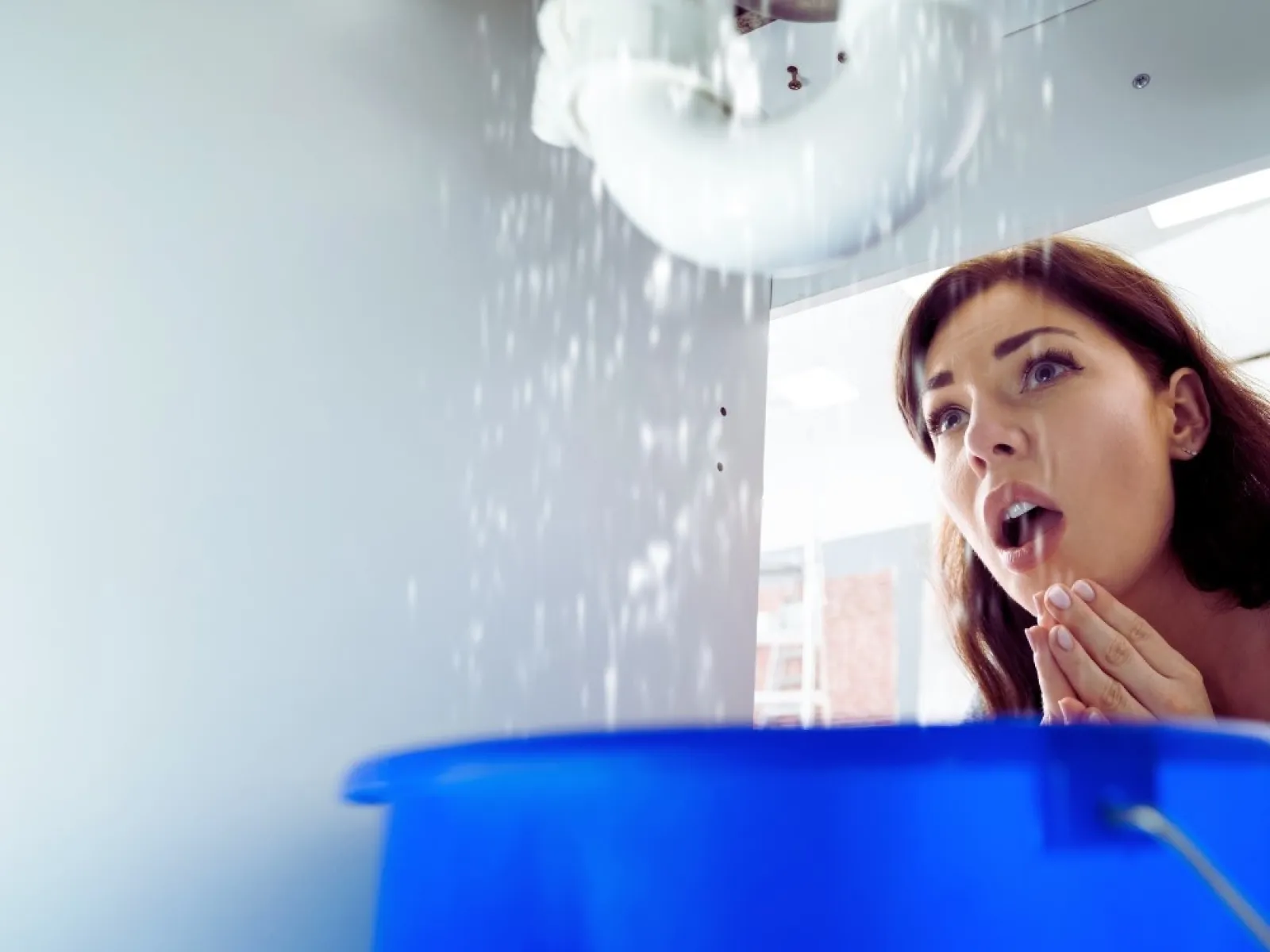 Woman looking shocked at water leaking from pipes into a blue bucket under a sink indoors.