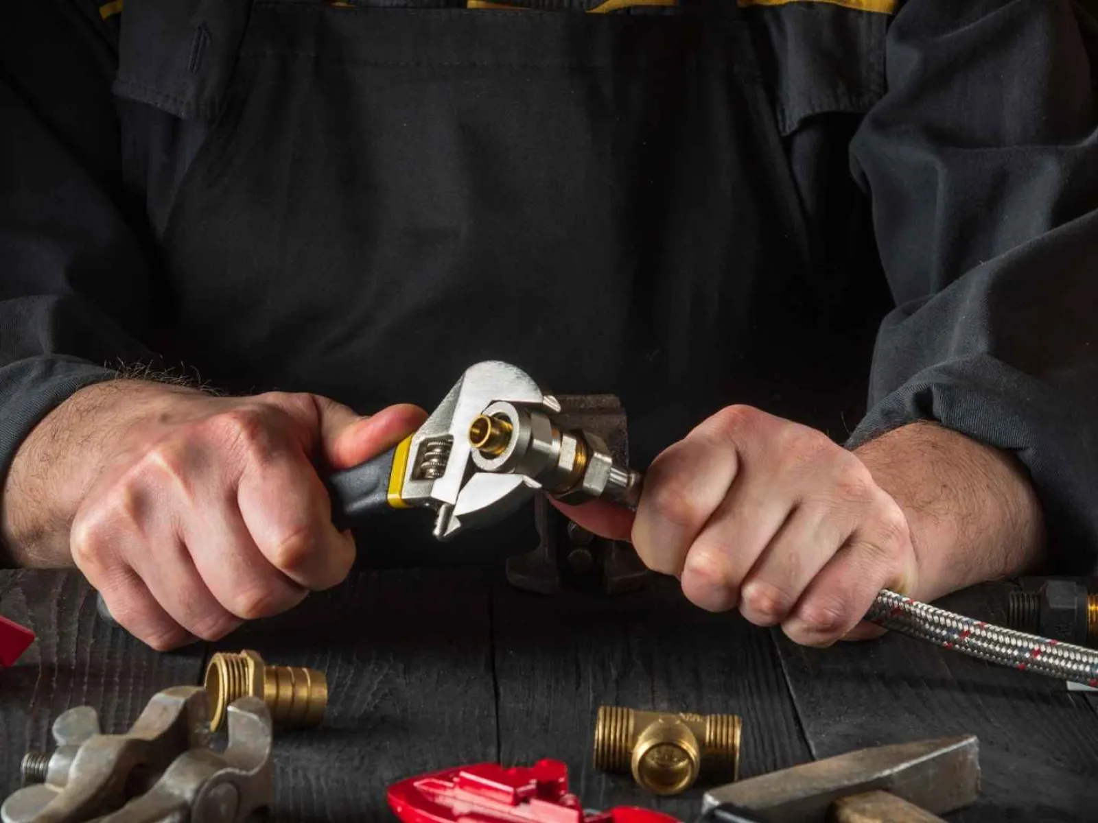 Hands of a plumber using a wrench to tighten a metal hose fitting on a dark workbench with plumbing tools.