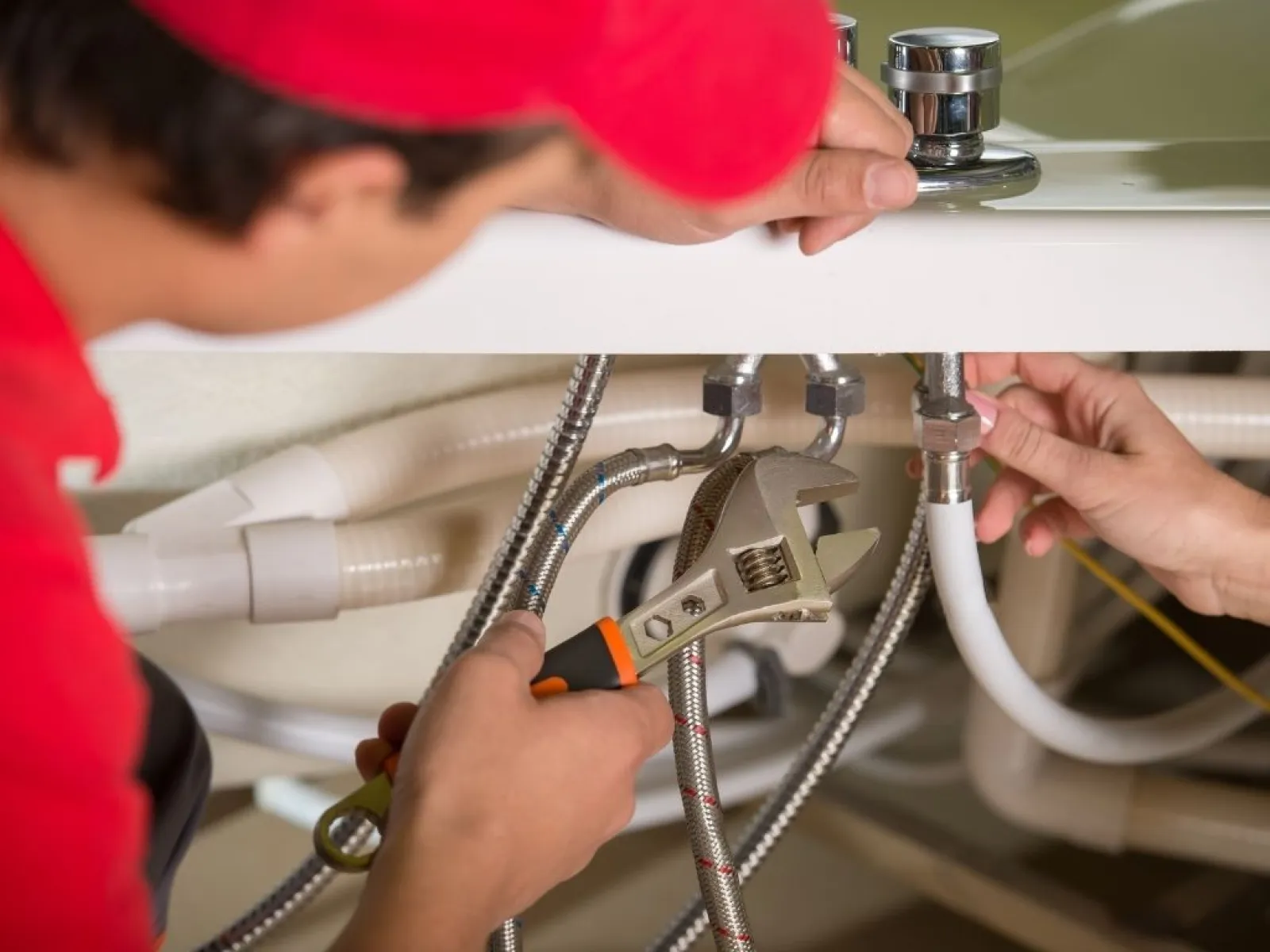 Plumber fixing kitchen sink pipes with adjustable wrench under the counter, hands working on fittings.