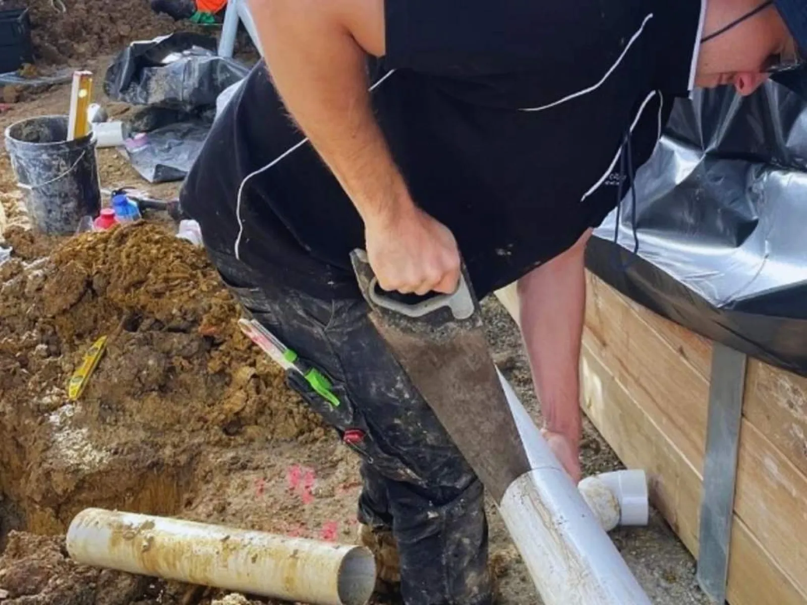 Worker cutting a white PVC pipe at a construction site with dirt and tools around.