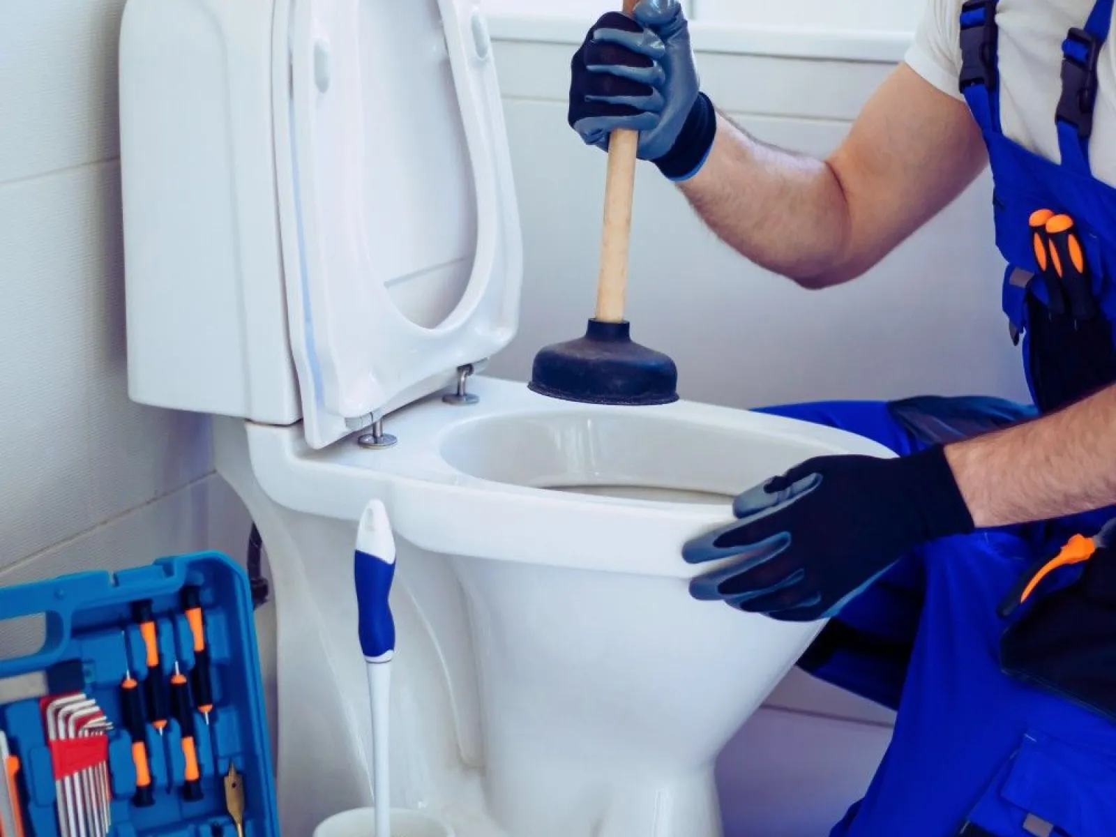 Plumber wearing gloves unclogs a white toilet with a plunger in a bathroom with tools nearby