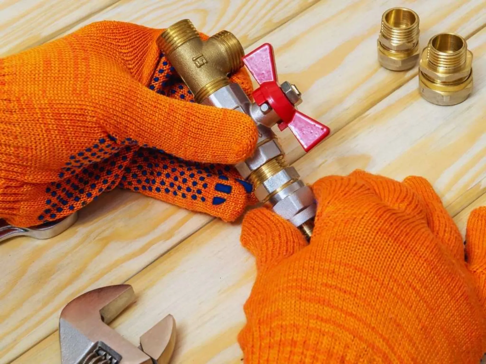 Hands in orange gloves holding a brass valve with a red handle on a wooden surface with tools nearby