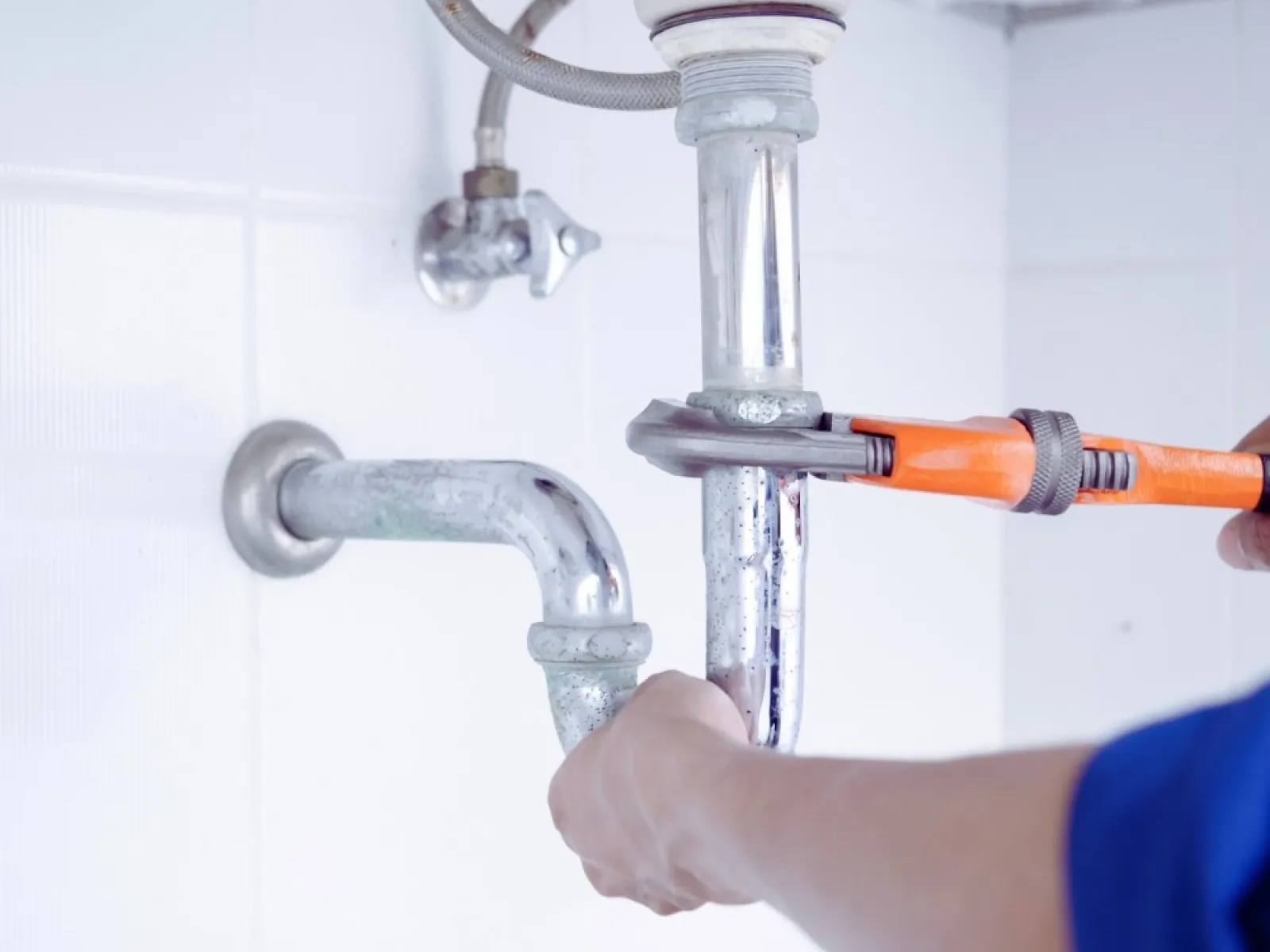 Plumber fixing a white sink pipe with an orange wrench in a tiled bathroom wall setting.