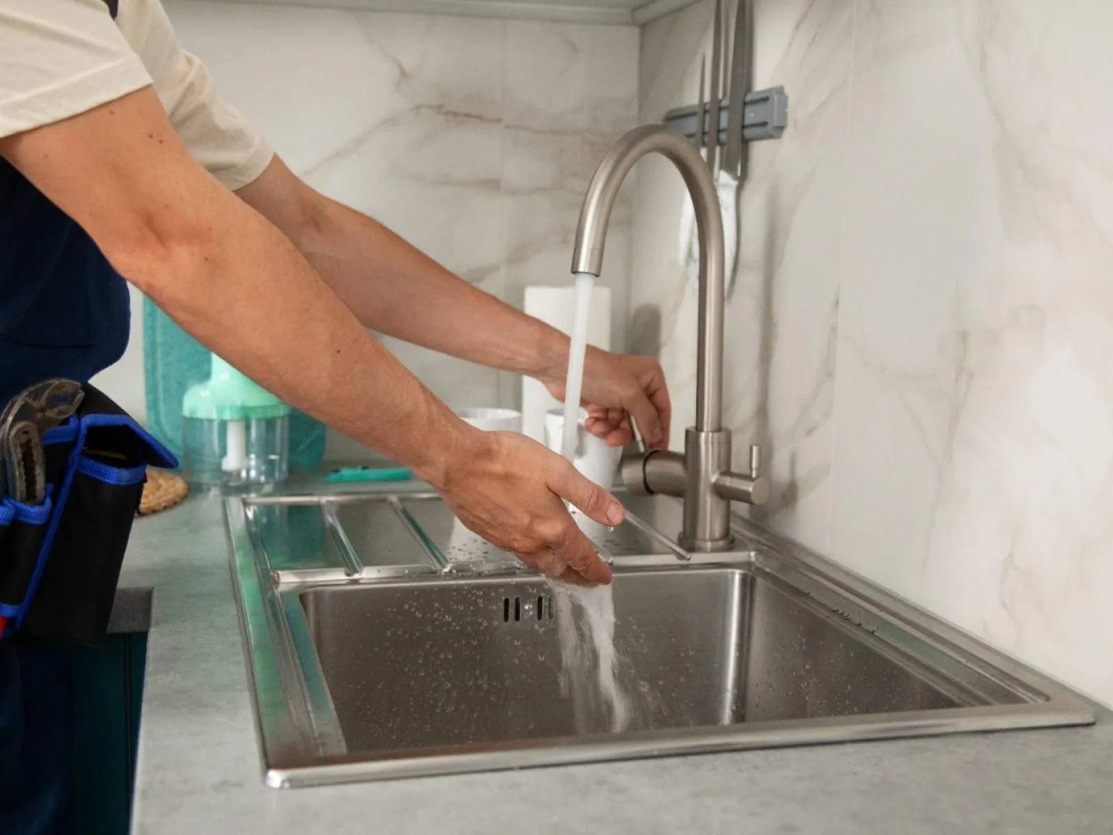 Plumber in work uniform running water from a faucet into a stainless steel kitchen sink with tools in belt.