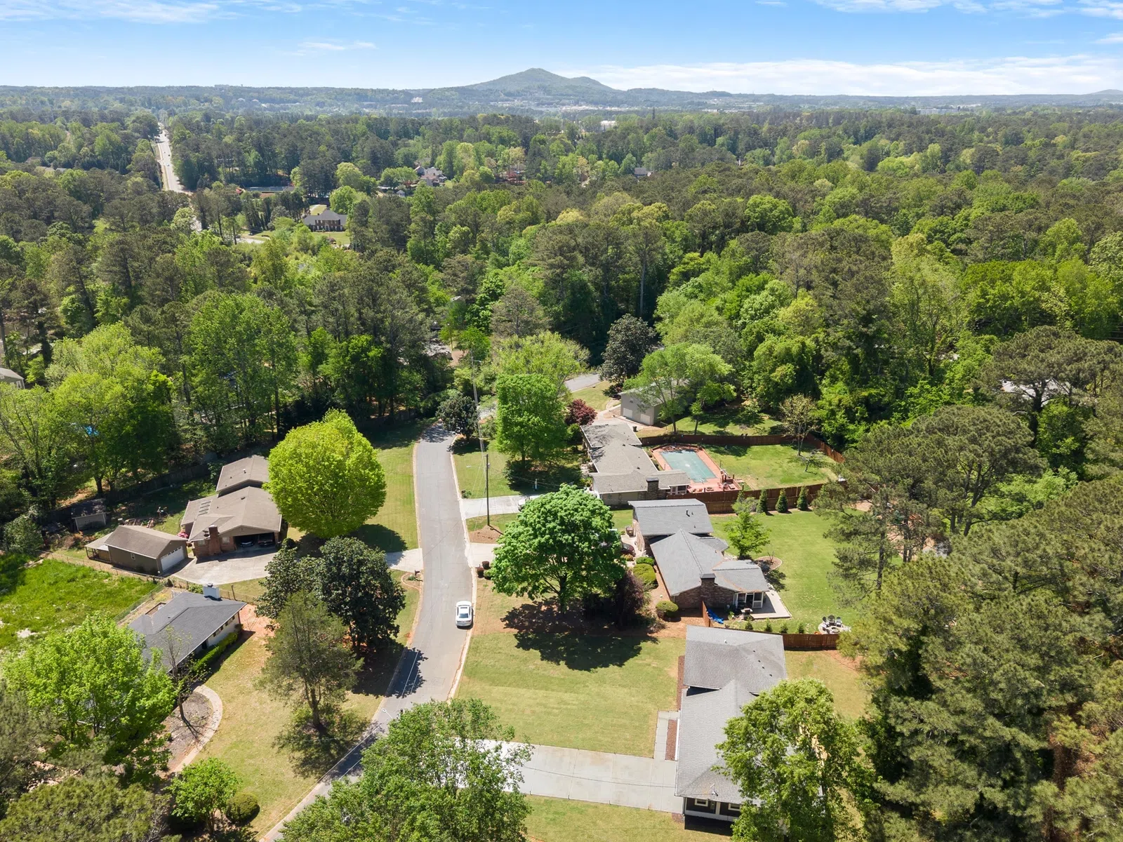 Aerial view of a suburban neighborhood with houses, green trees, lawns, and a distant mountain under a clear blue sky.