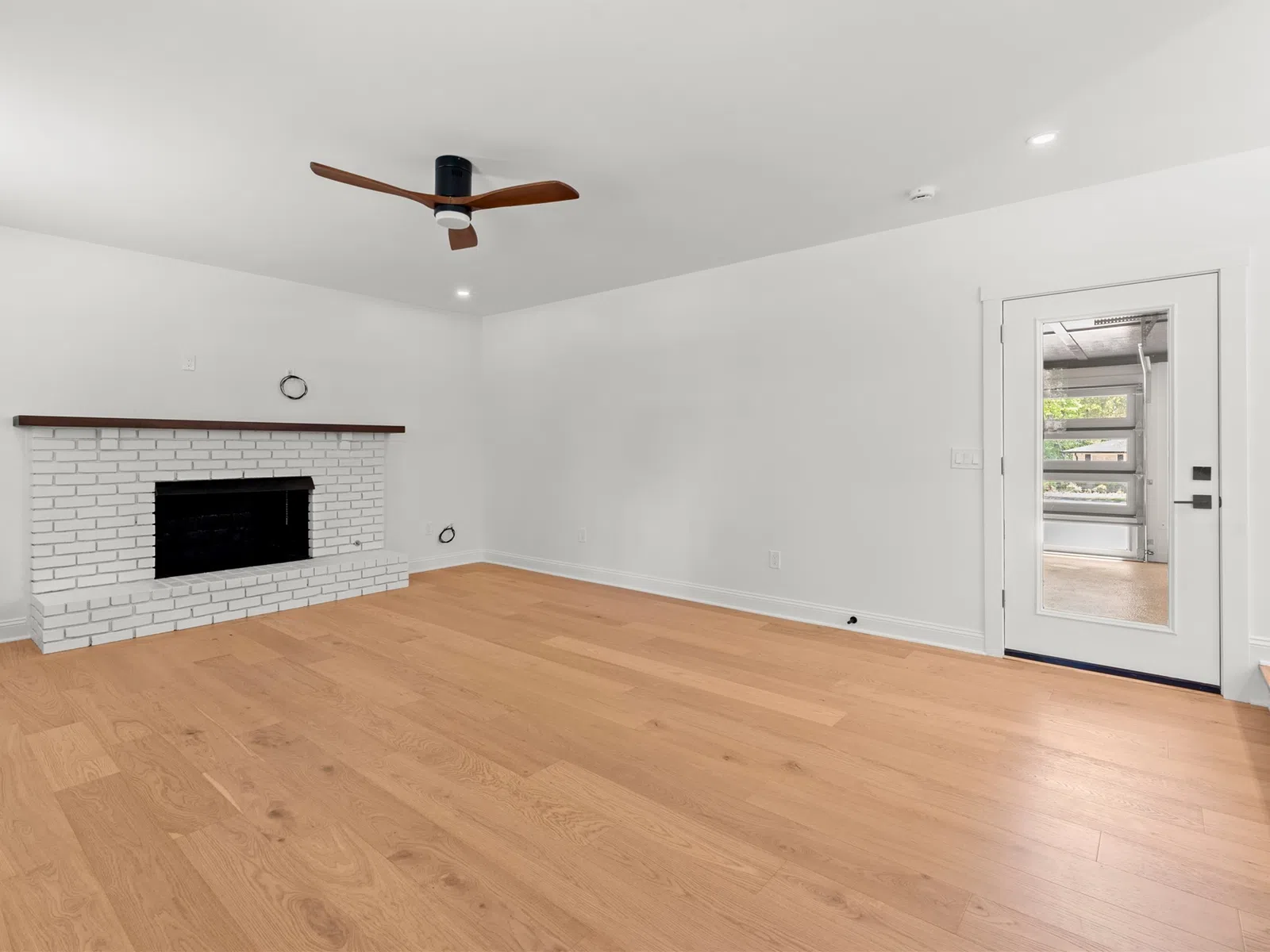 Empty modern living room with light hardwood floors, white walls, brick fireplace, ceiling fan, and glass door.
