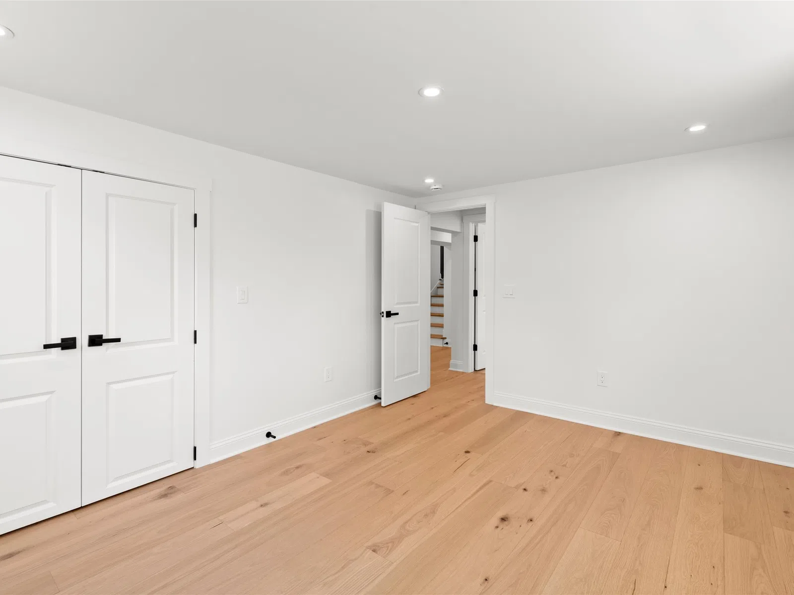 Empty white room with light wood flooring, white doors, and recessed ceiling lights in a modern home.