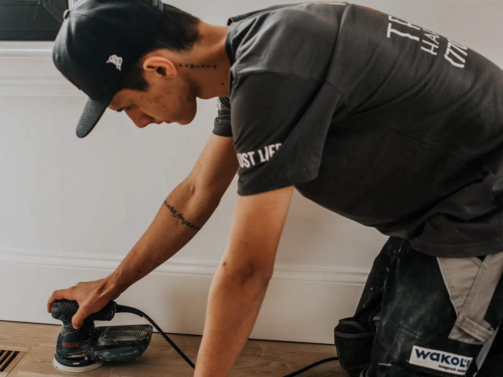 Man in cap sanding wooden floor near white wall using electric sander powered by Bosch