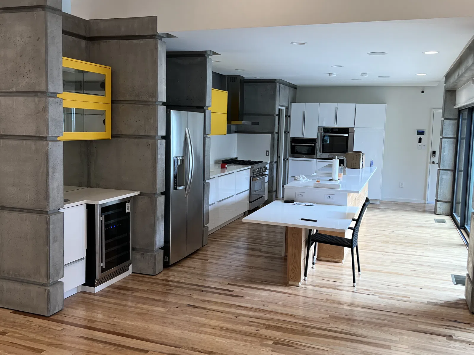 Modern kitchen with hardwood floors, concrete pillars, yellow accents, stainless steel appliances, and white cabinetry.