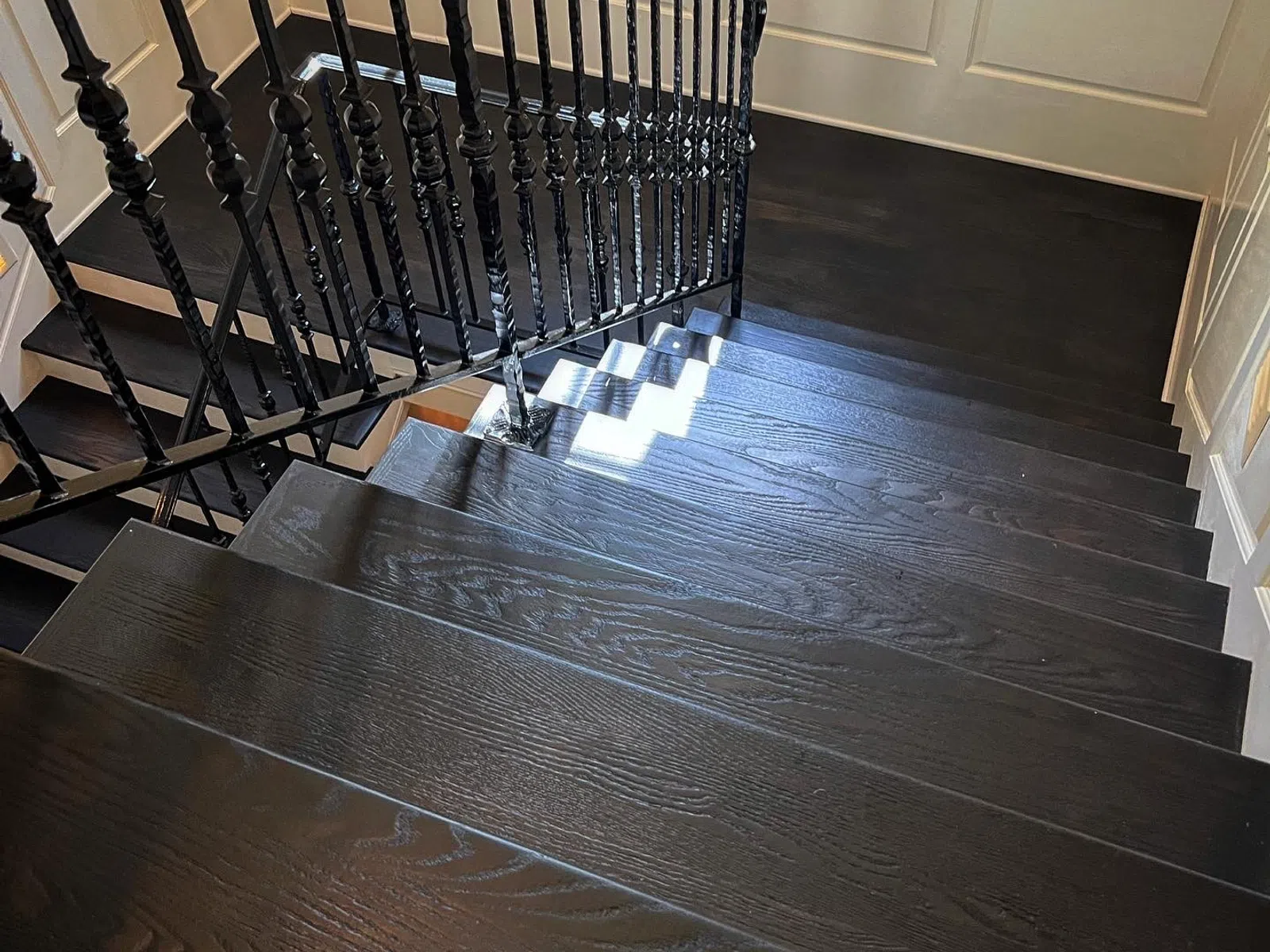 Dark wooden staircase with black iron balusters in a well-lit interior space with white paneled walls.