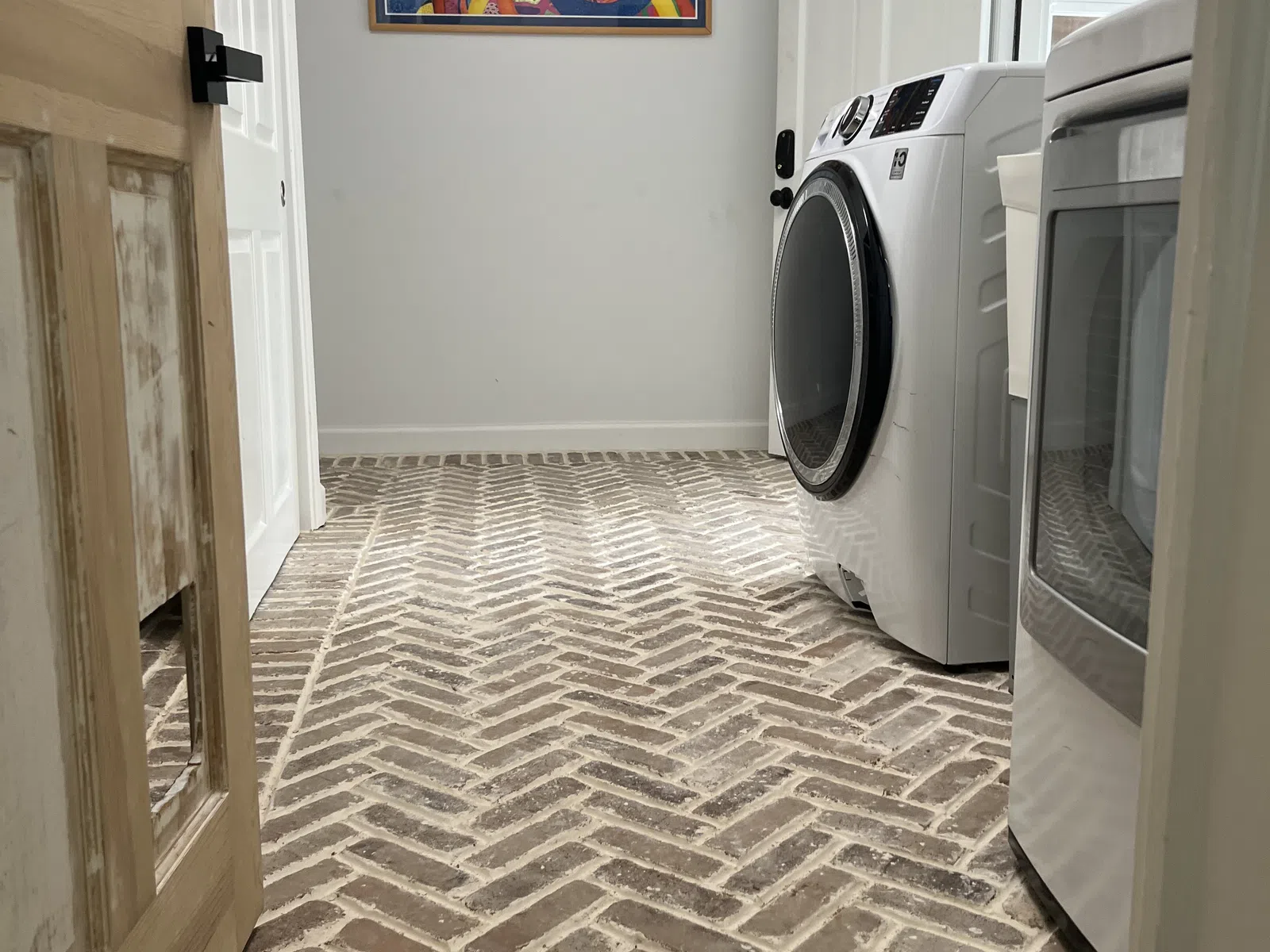 Laundry room with herringbone brick flooring, white washer and dryer, wooden door, and colorful abstract wall art.