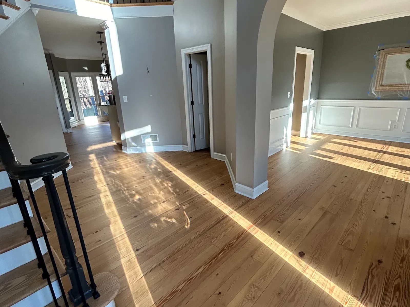 Sunlit empty room with wooden floors, beige walls, arched doorway, and black railing staircase in modern home.