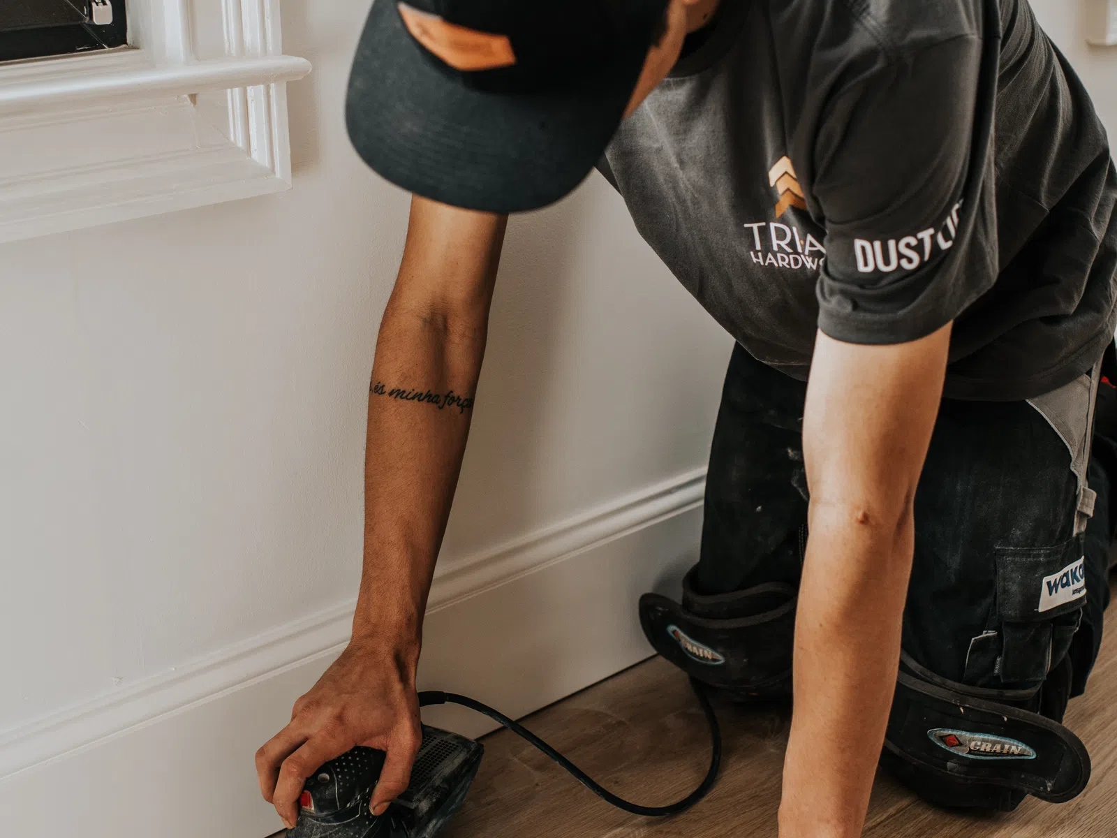 Worker wearing black cap and shirt sanding wooden floor near white wall with power sander