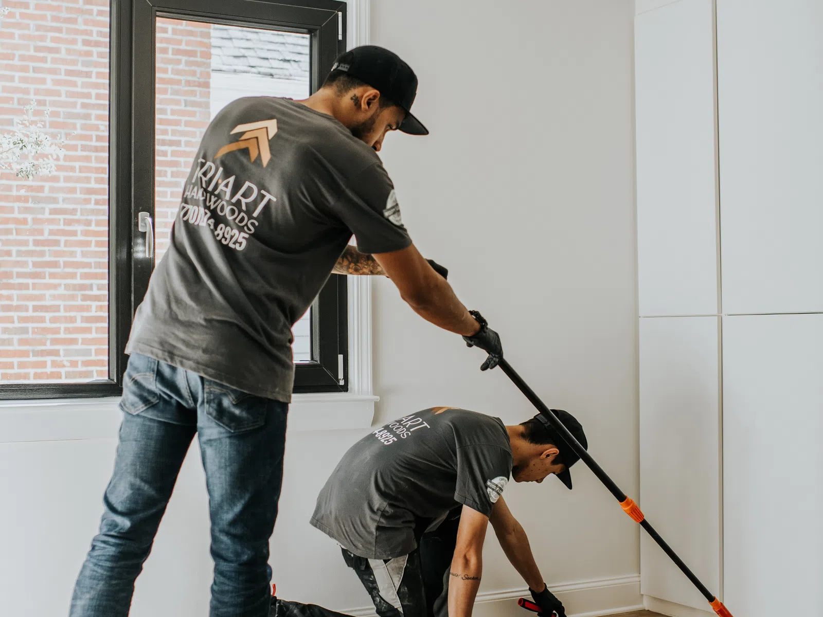 Two workers installing or finishing a hardwood floor in a bright, modern room with large window and white walls.