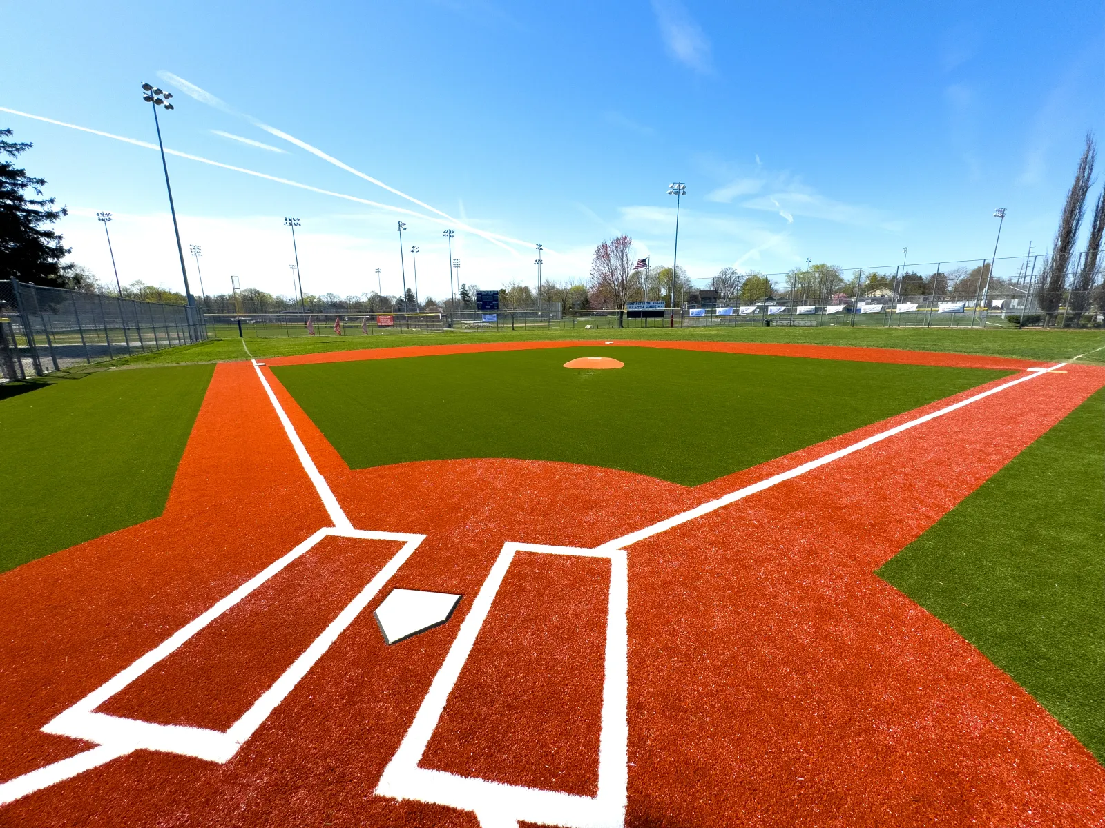 Empty baseball field with green artificial turf and bright red infield under a clear blue sky.