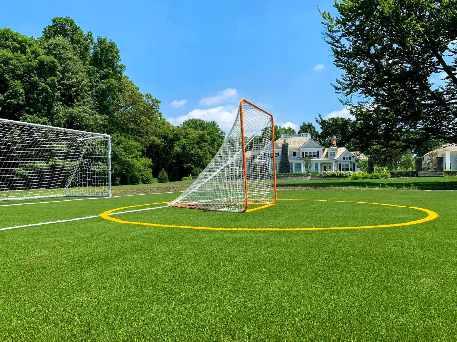 Lacrosse and soccer goals on a green field with trees and residential houses under a clear blue sky.