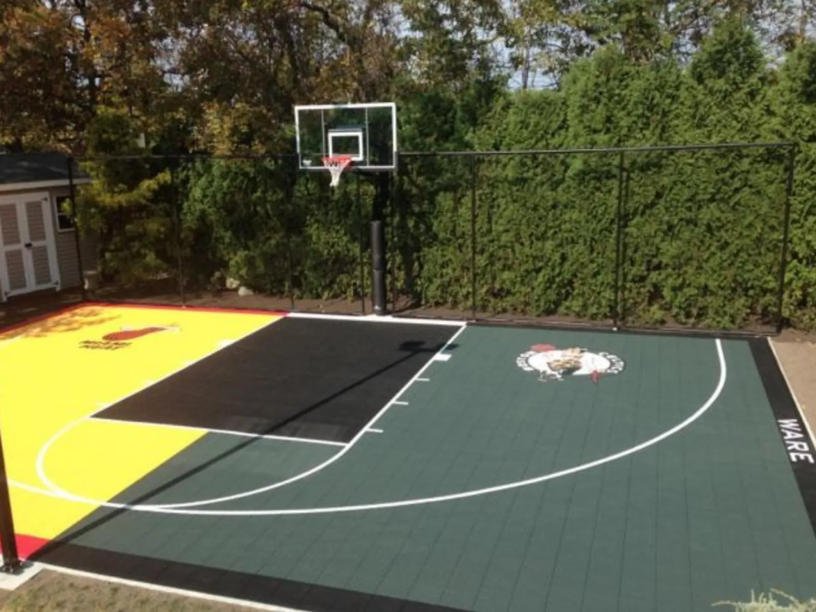 Outdoor residential basketball court with green, yellow, and black colored sections, featuring a hoop and logos.
