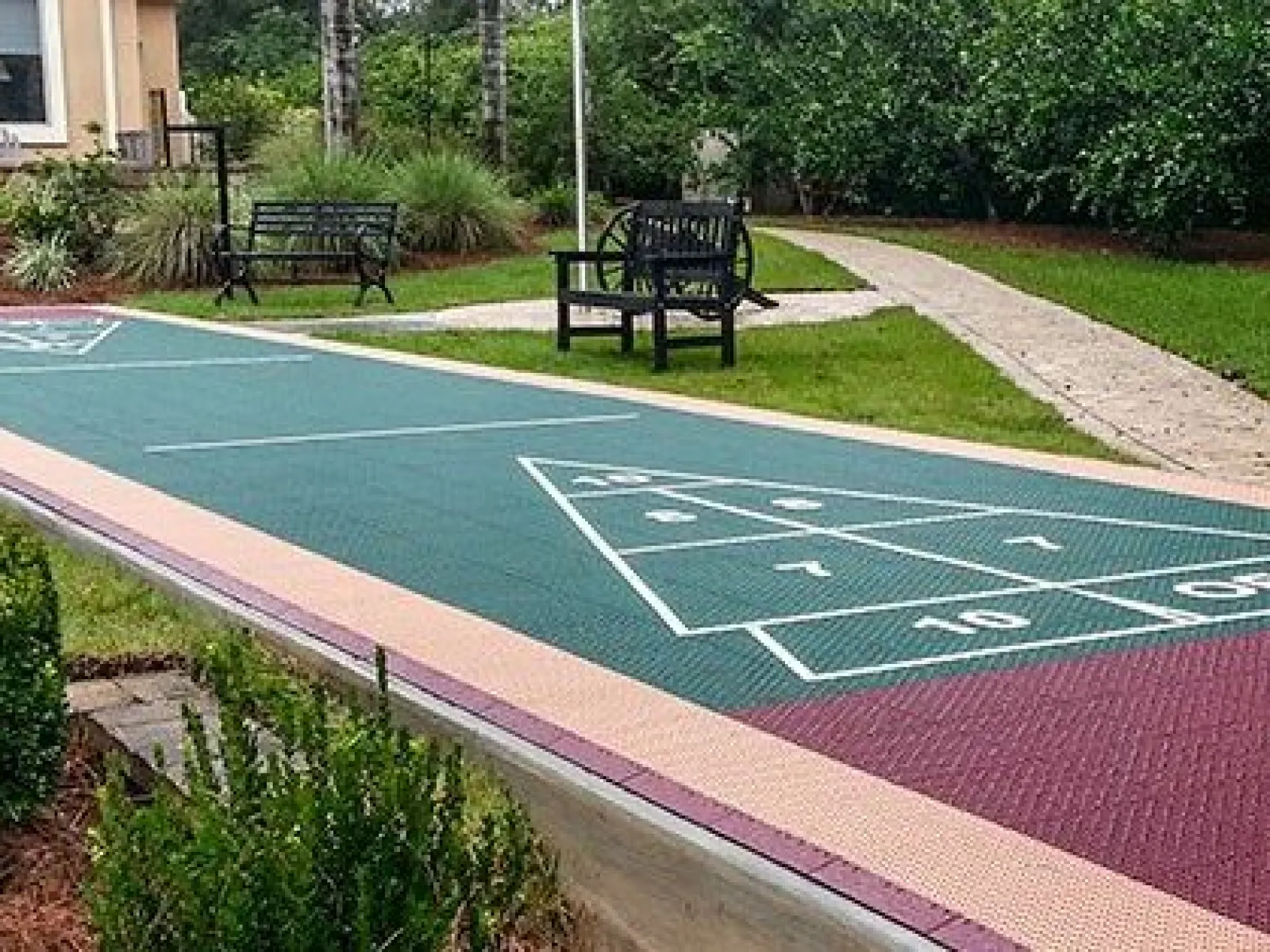 Outdoor shuffleboard court with green playing surface, benches, and surrounding greenery in a park setting