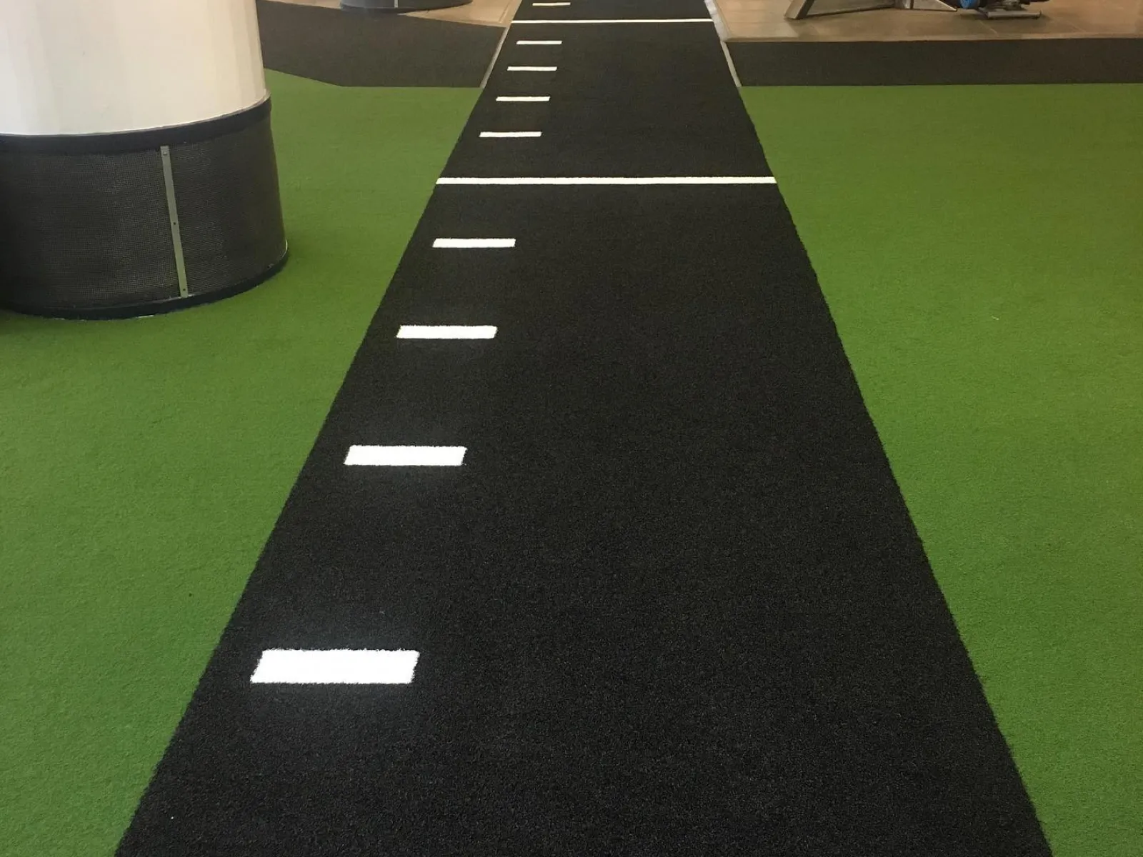 Black turf running track with white markings inside a gym surrounded by green flooring and equipment.