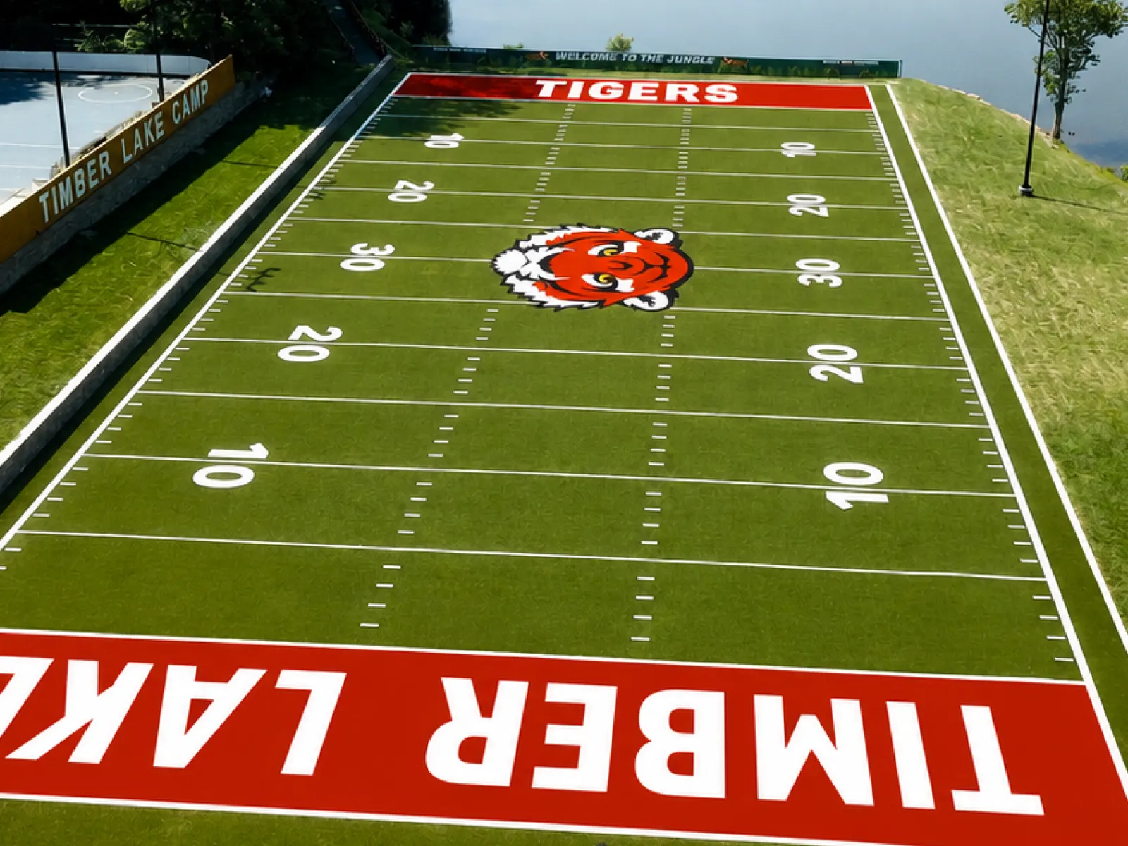 Aerial view of Timber Lake football field with Tigers logo beside a lake and surrounding trees under clear sky.