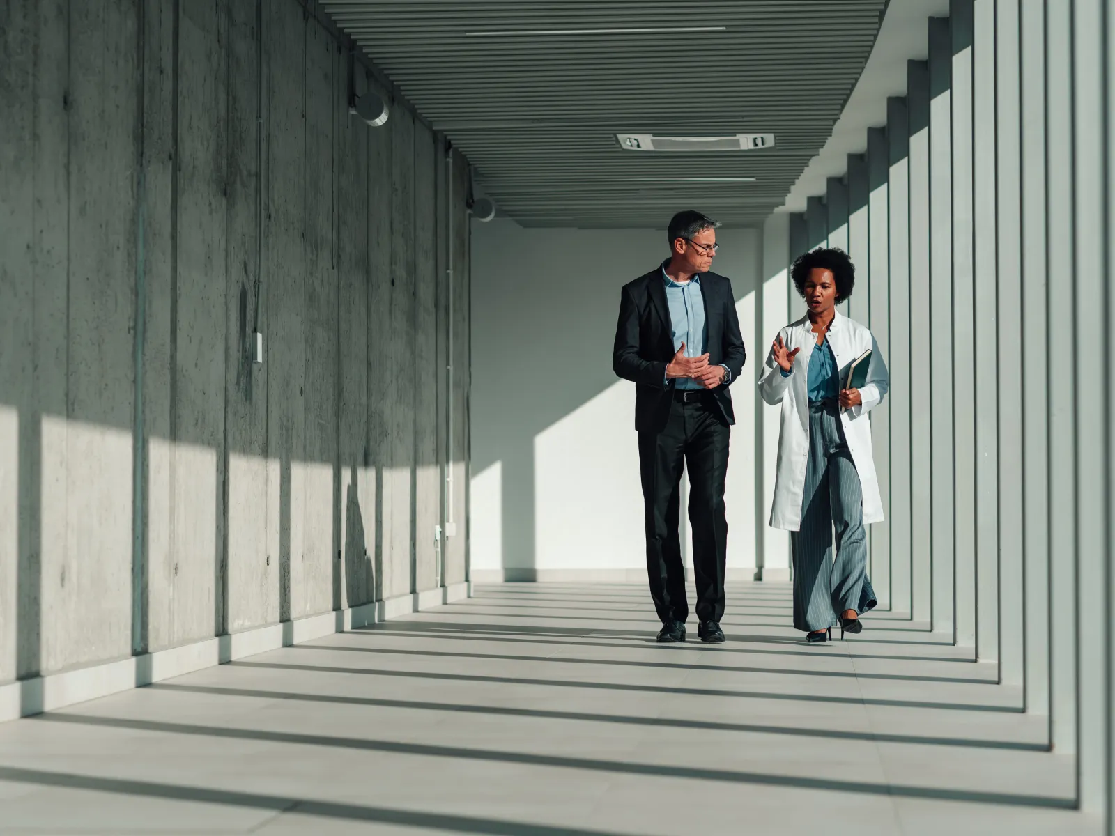 Two professionals walking and discussing in a modern corridor with glass walls and strong natural light.
