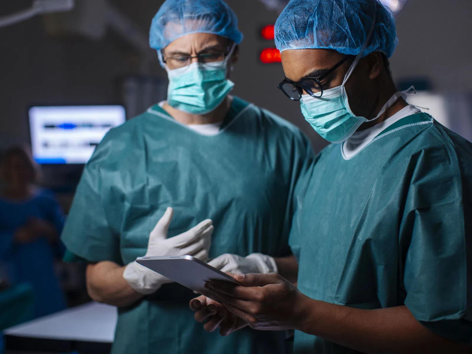 Two surgeons wearing scrubs, masks, and caps review patient information on a tablet in an operating room.