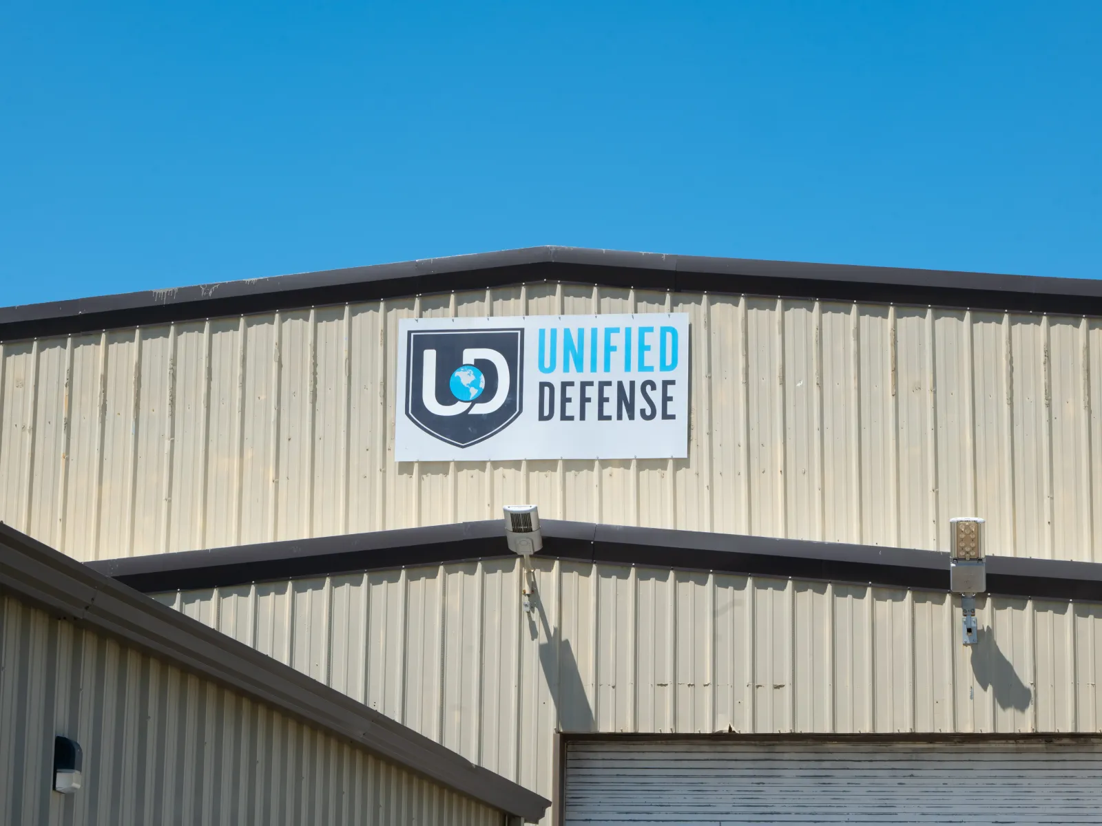 Unified Defense logo on a beige industrial building under a clear blue sky.