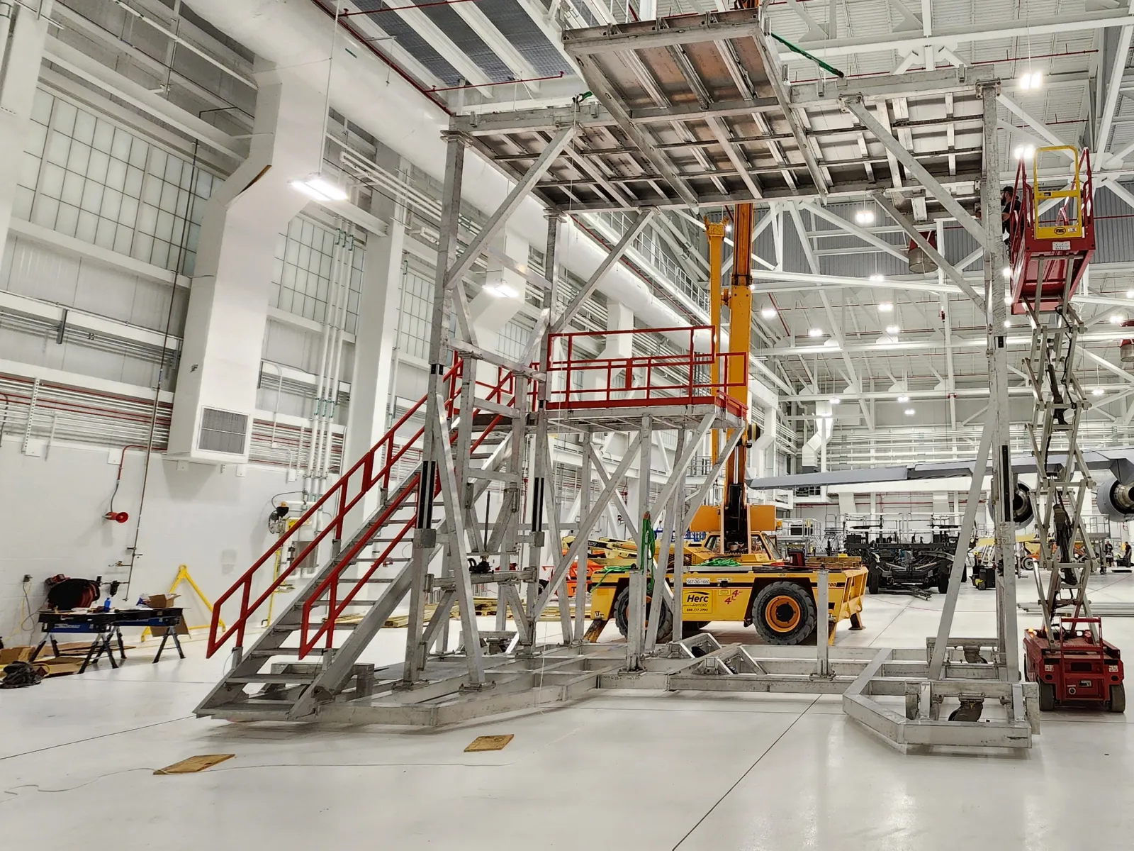 Aircraft maintenance hangar with scaffolding, platforms, forklifts, and a military jet under bright industrial lighting.