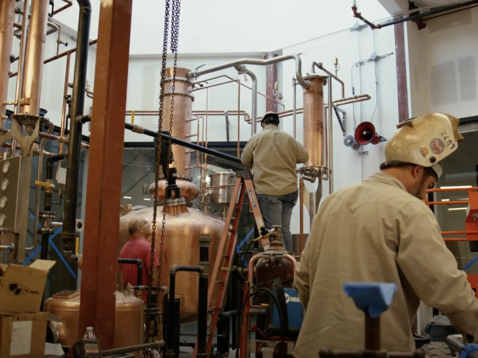 Workers operate copper distillation equipment inside an industrial distillery with pipes and machinery.