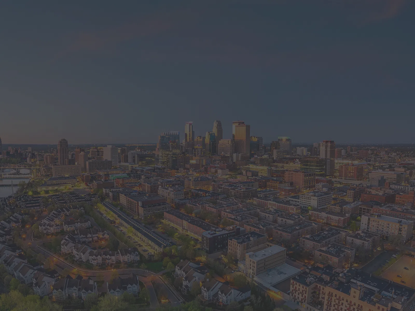Aerial night view of a city skyline with illuminated buildings and streets under a darkening sky.