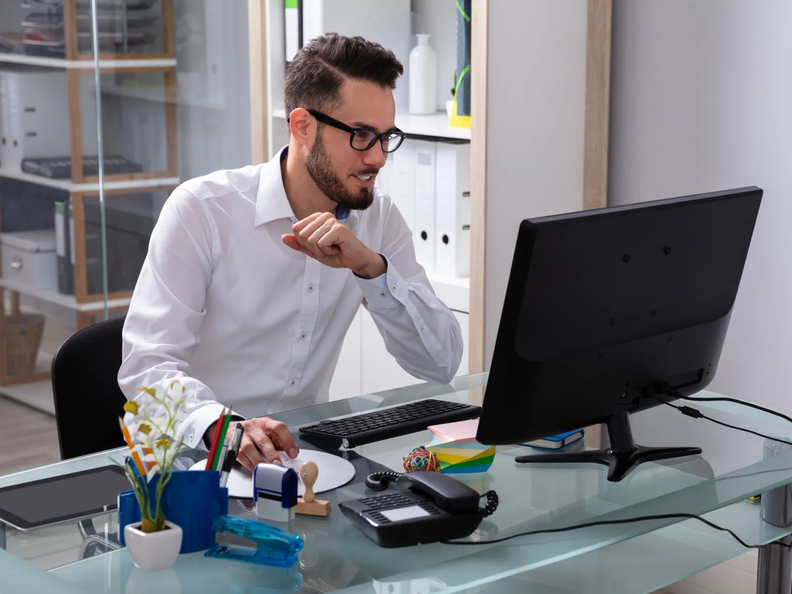 Man in glasses working on desktop computer at modern office desk with office supplies and decor.