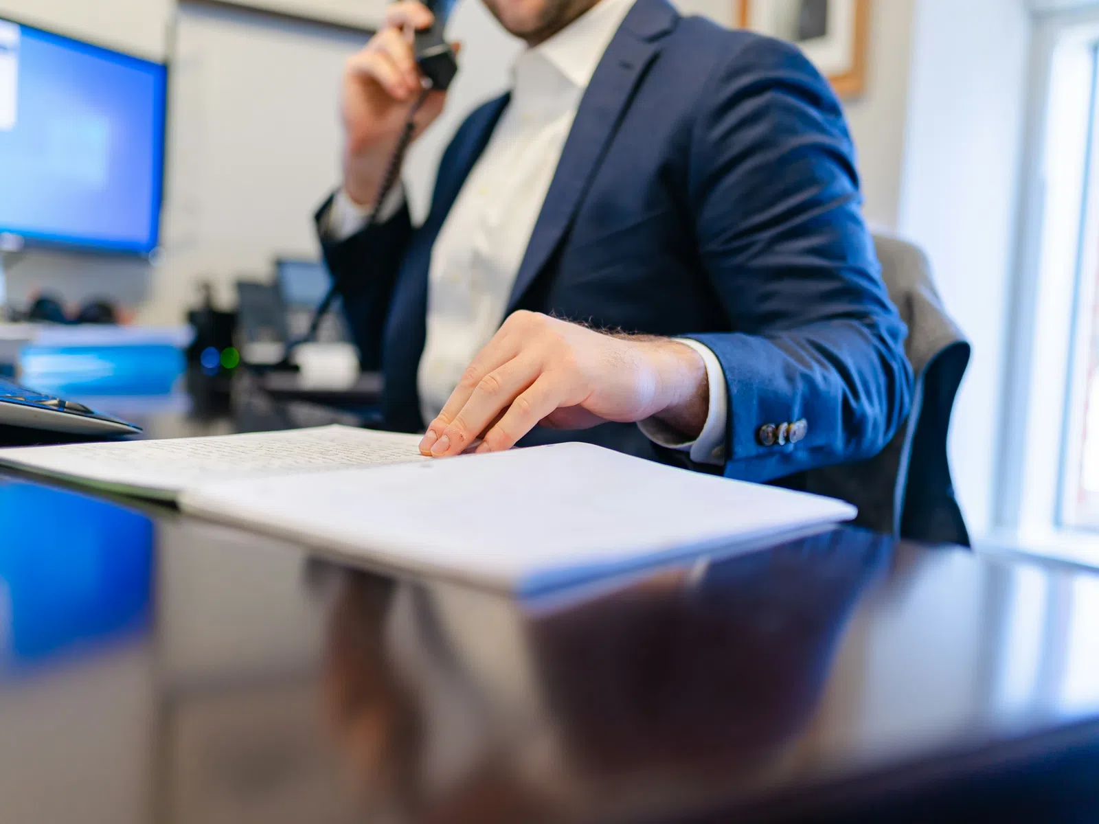 Businessman in blue suit talking on phone and reviewing documents at office desk with computer.
