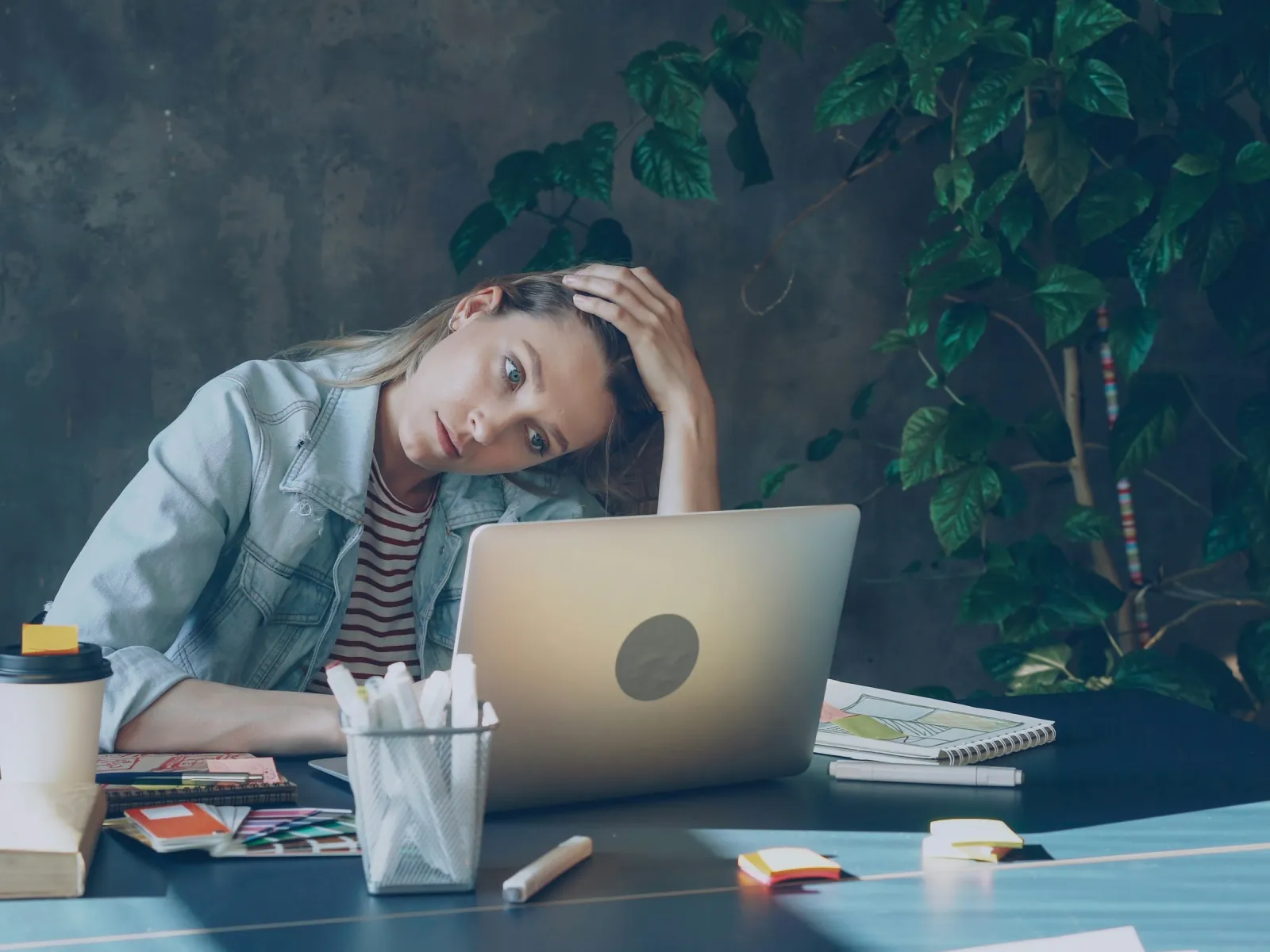 Young woman in denim jacket appears stressed working on laptop at a desk with plants and office supplies.