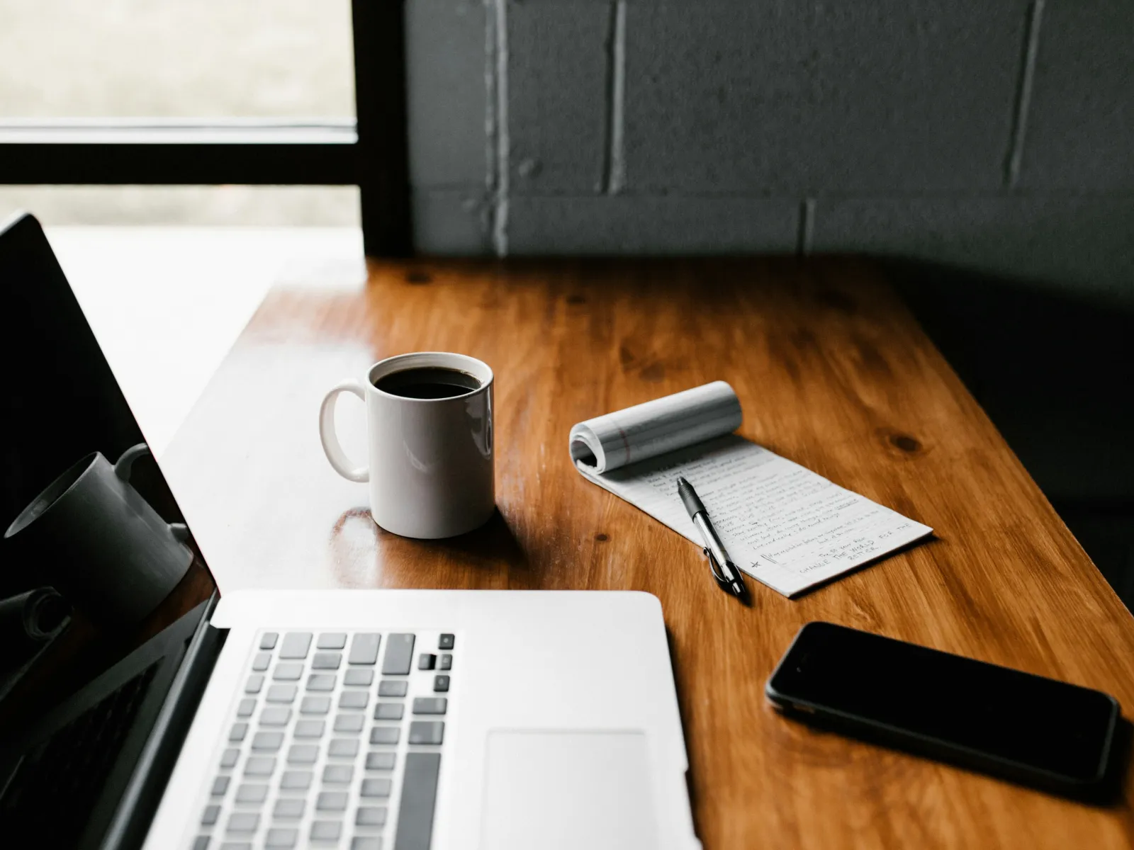 Laptop, coffee mug, notepad with pen, and smartphone on wooden desk near window with natural light.