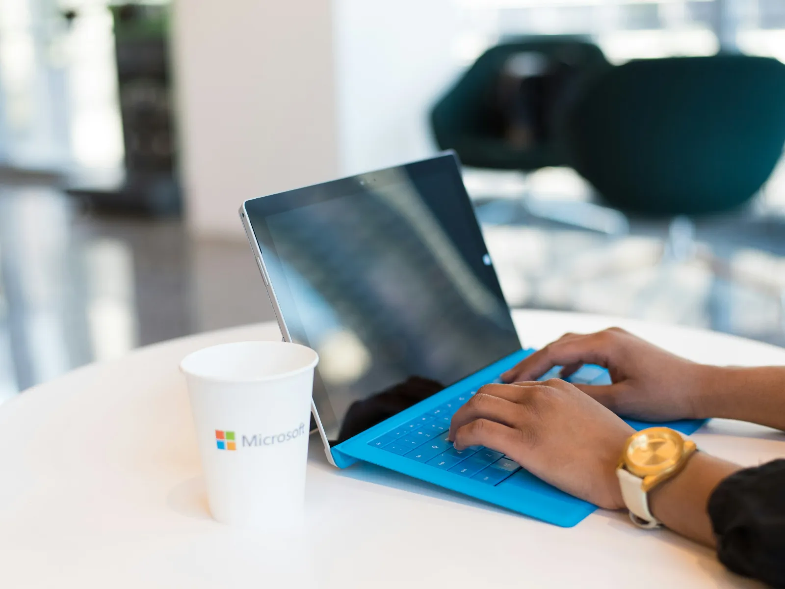 Hands typing on a blue keyboard attached to a tablet on a white table with a Microsoft coffee cup nearby
