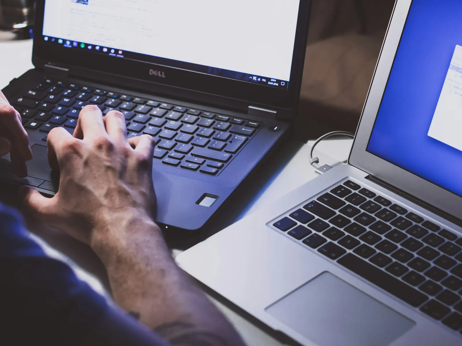 Person typing on Dell laptop beside an open MacBook Air on a white table in a workspace.