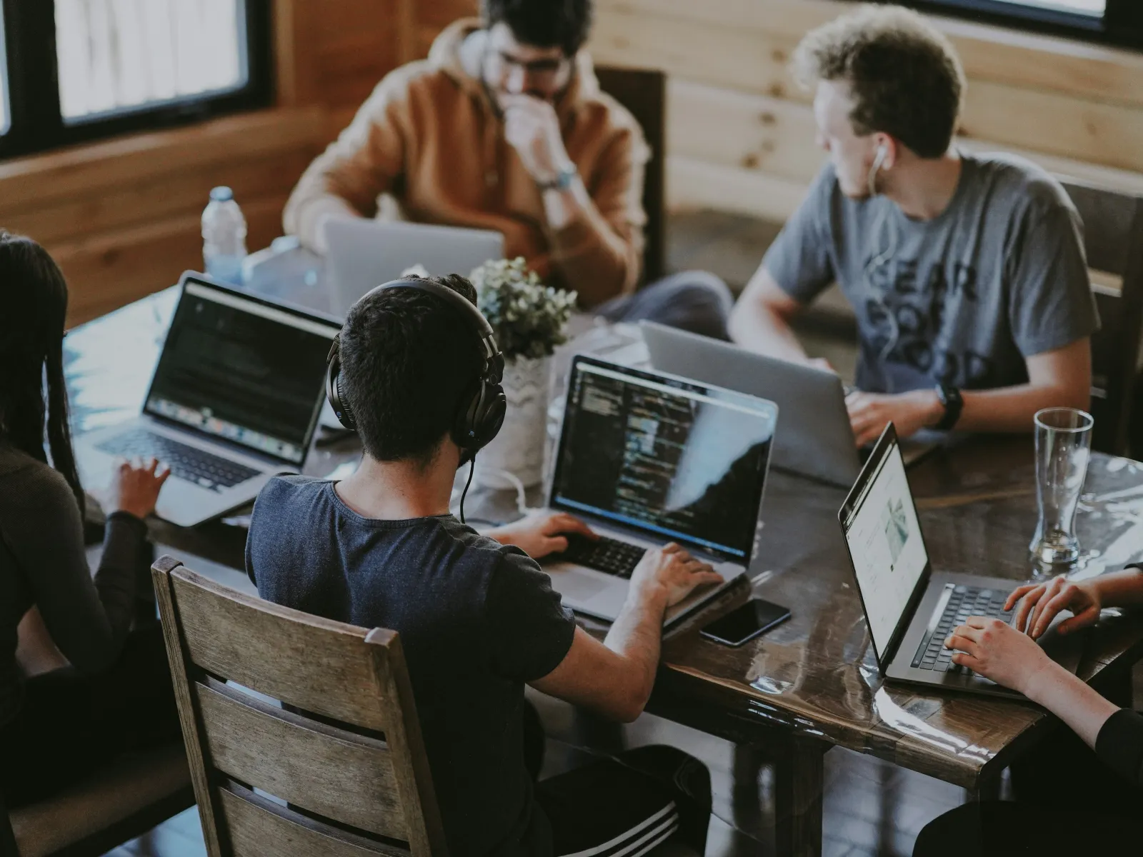 Young professionals collaborating and coding on laptops around a wooden table in a cozy modern office.