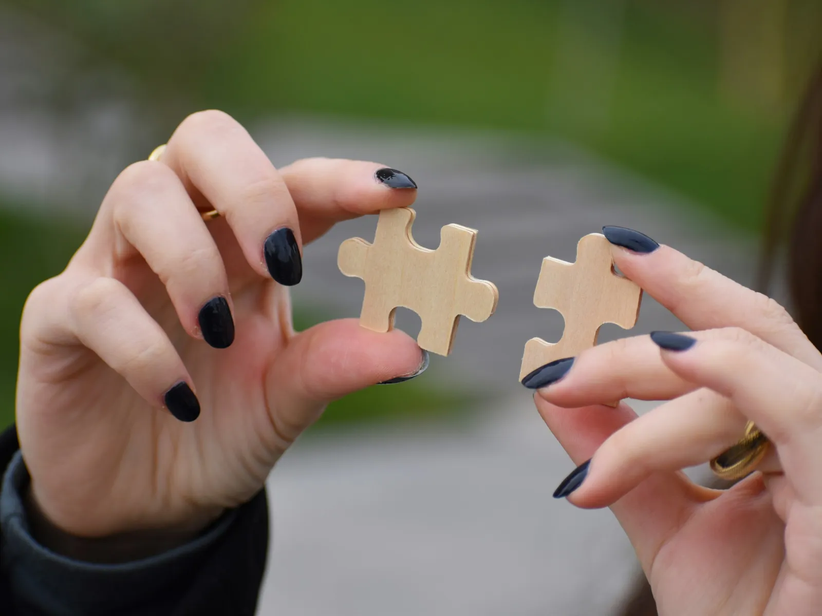 Hands with black nail polish holding two wooden puzzle pieces about to connect outdoors.