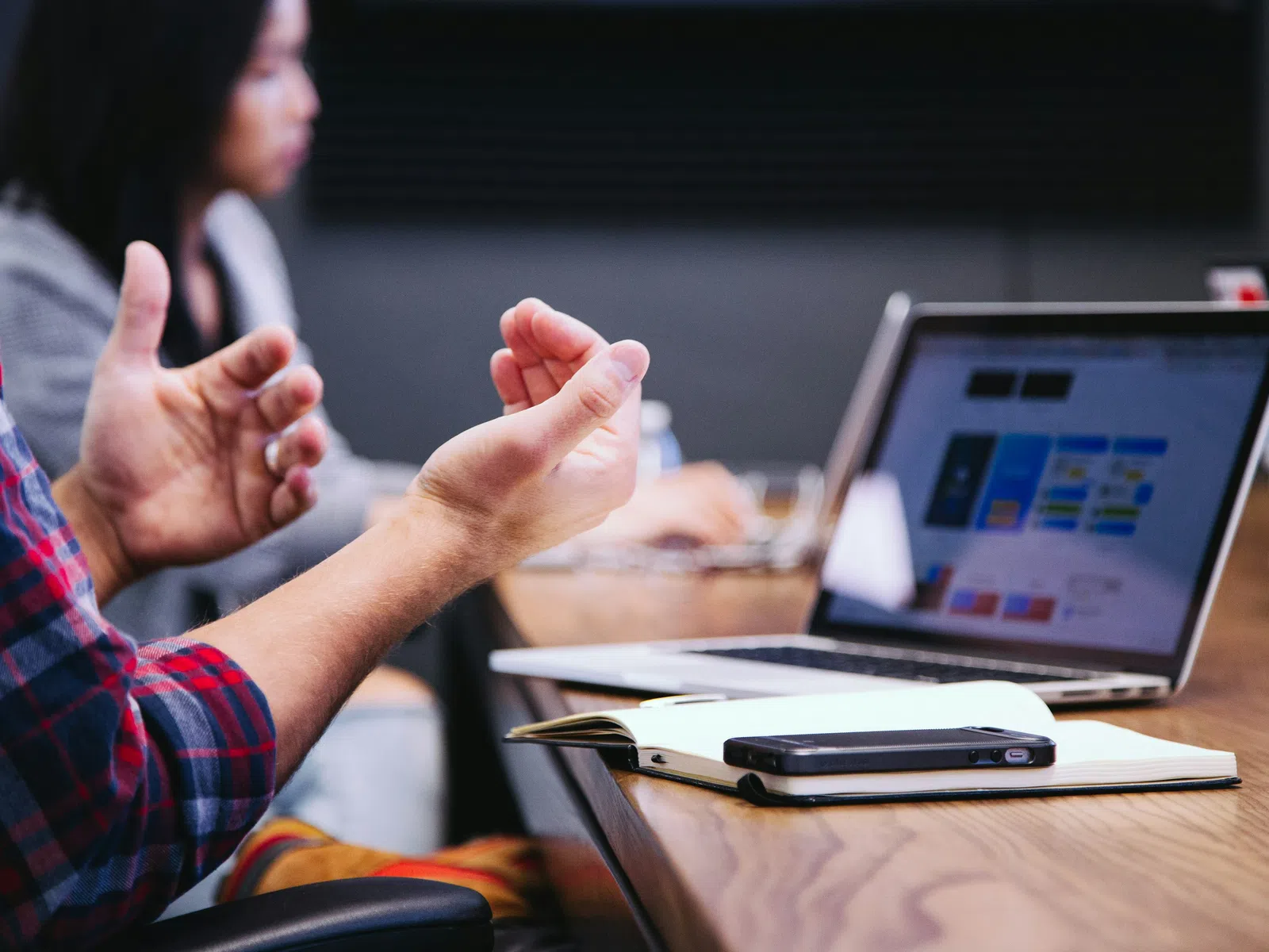 Person gesturing with hands during a meeting with laptop, notebook, and smartphone on the wooden table.