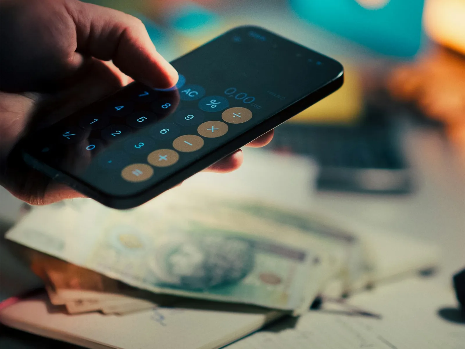 Hand holding smartphone with calculator app open over notebook and scattered currency notes on a desk.