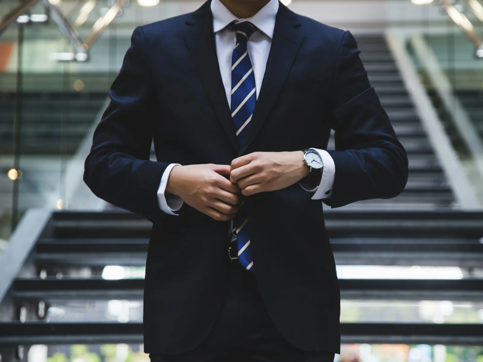 Man in navy suit adjusting jacket in front of modern staircase with glass railings.