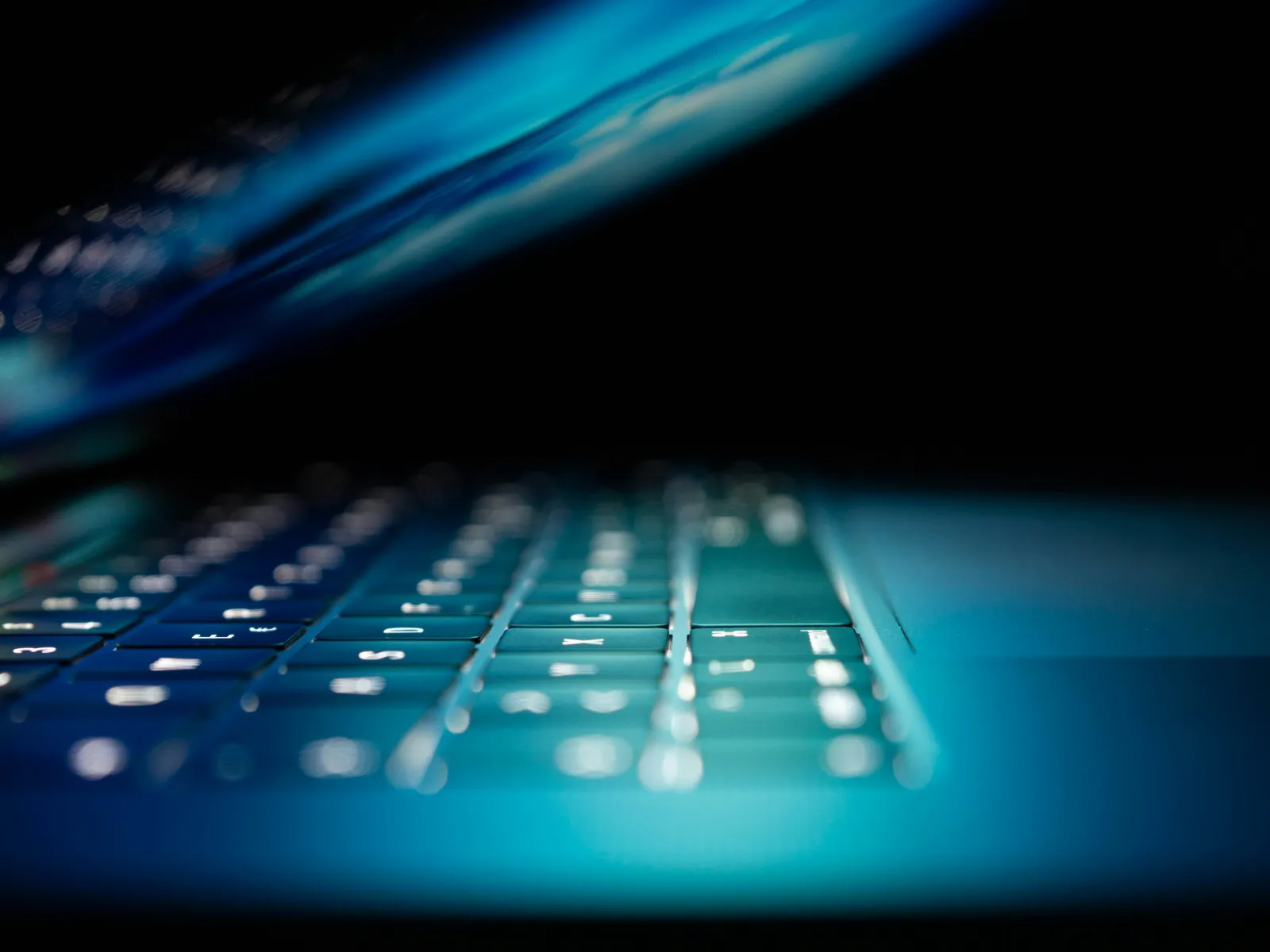 Close-up of a laptop keyboard with soft blue lighting and reflective screen in a dark environment.