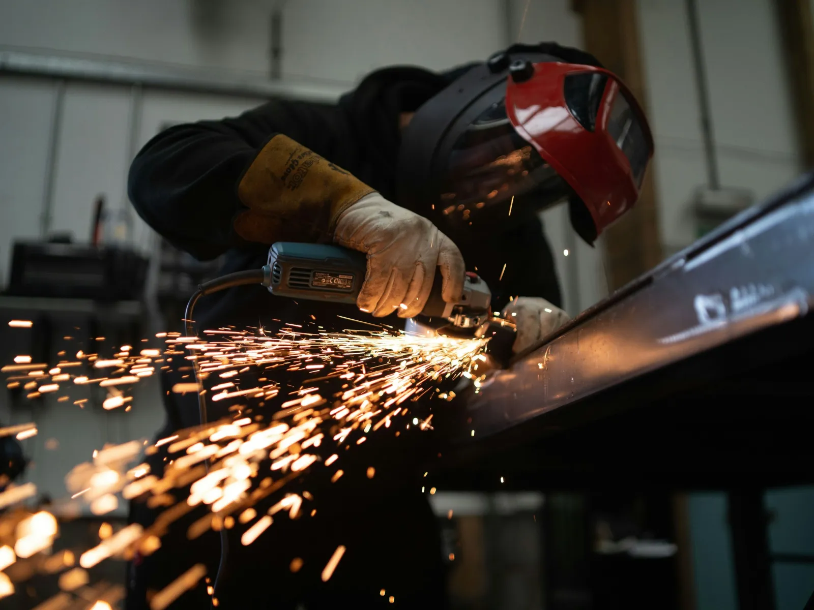 Worker wearing protective gear grinding metal, producing bright sparks in an industrial workshop setting.