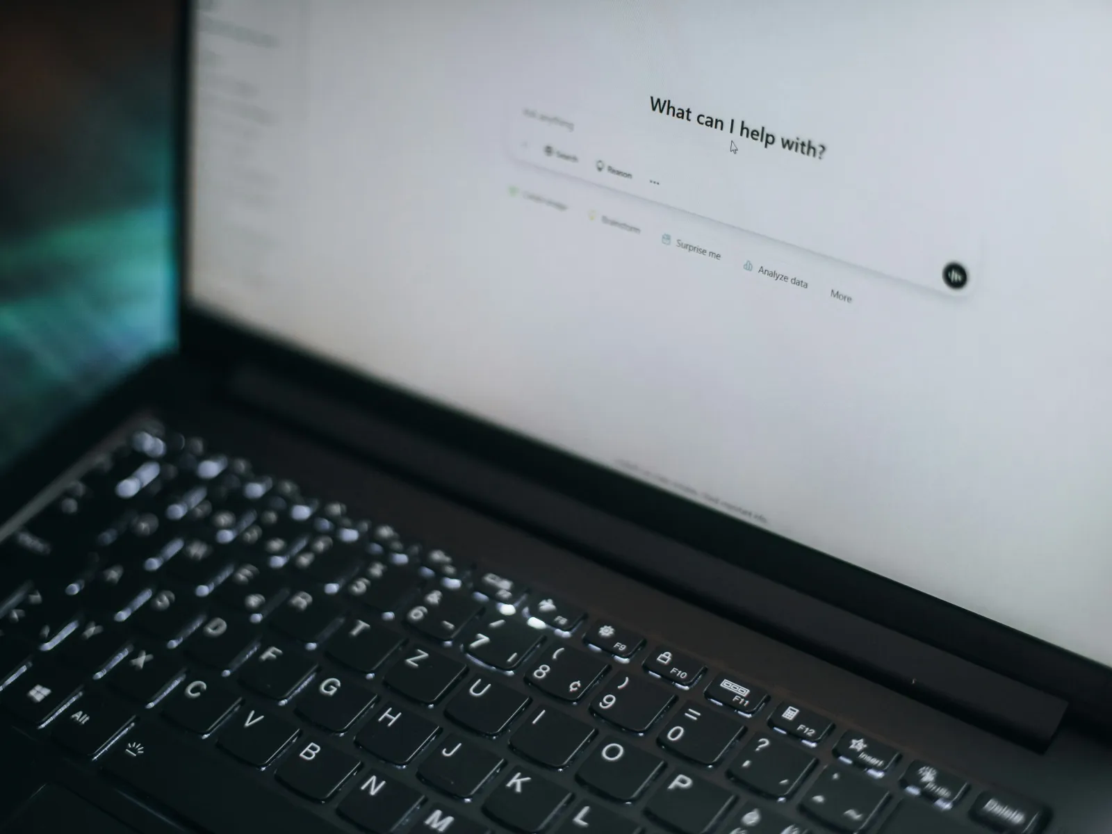Laptop keyboard and screen displaying a search bar with the text What can I help with?