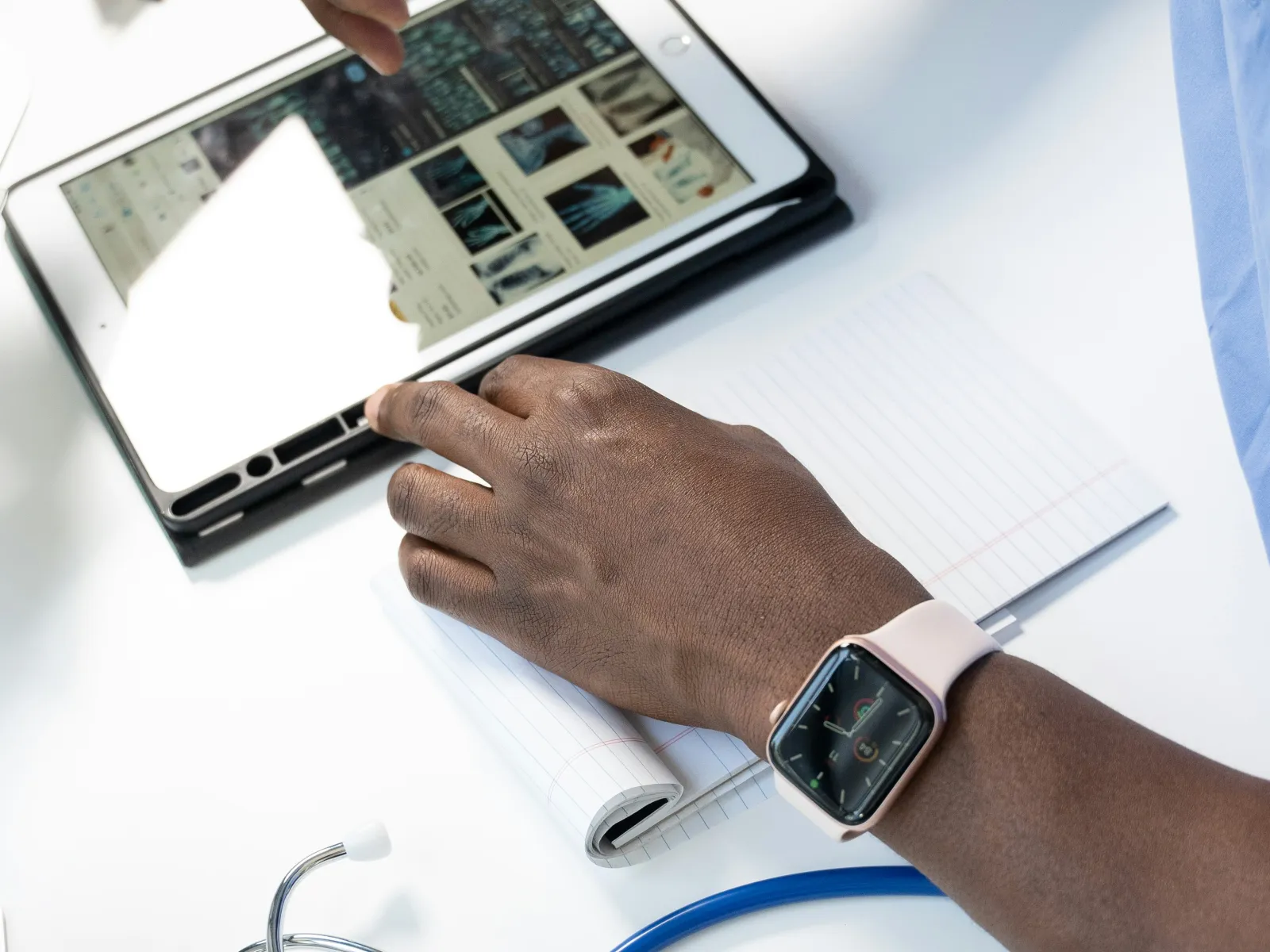 Person wearing smartwatch using tablet to view medical scans with stethoscope and notebook on desk