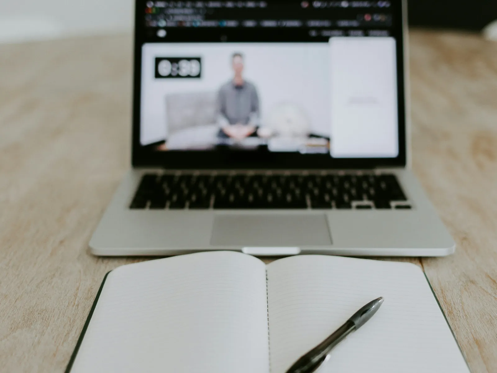 Open notebook with a pen on a wooden table in front of a laptop showing a blurred online video call.